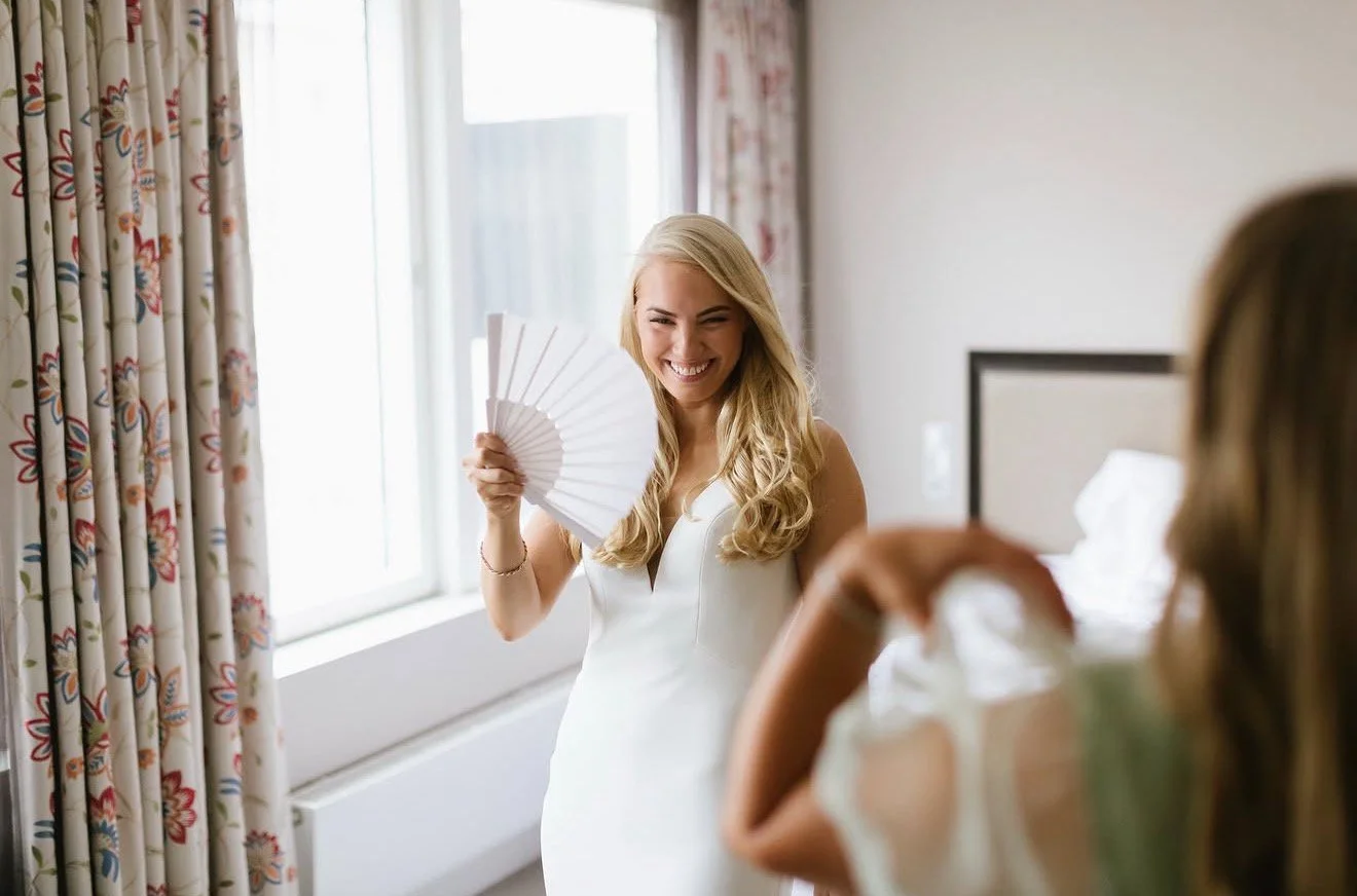 A blonde woman in a white dress smiling and holding a fan, interacting with another woman in a room with floral curtains and a bed in the background.