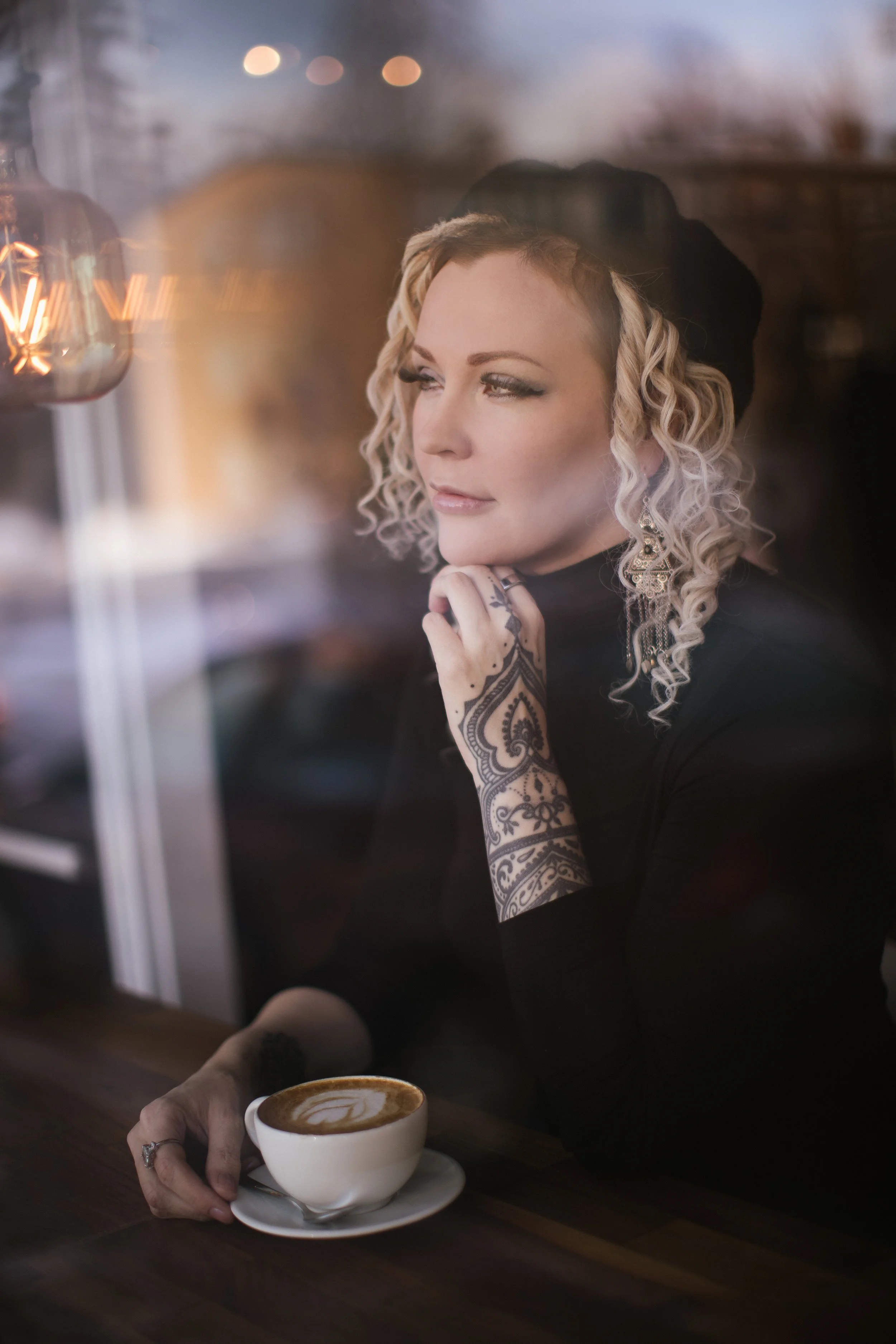 A woman with curly blonde hair, intricate earrings, and henna tattoos on her hand holds a cup of coffee with latte art at a coffee shop, seen through a window with warm light reflections.