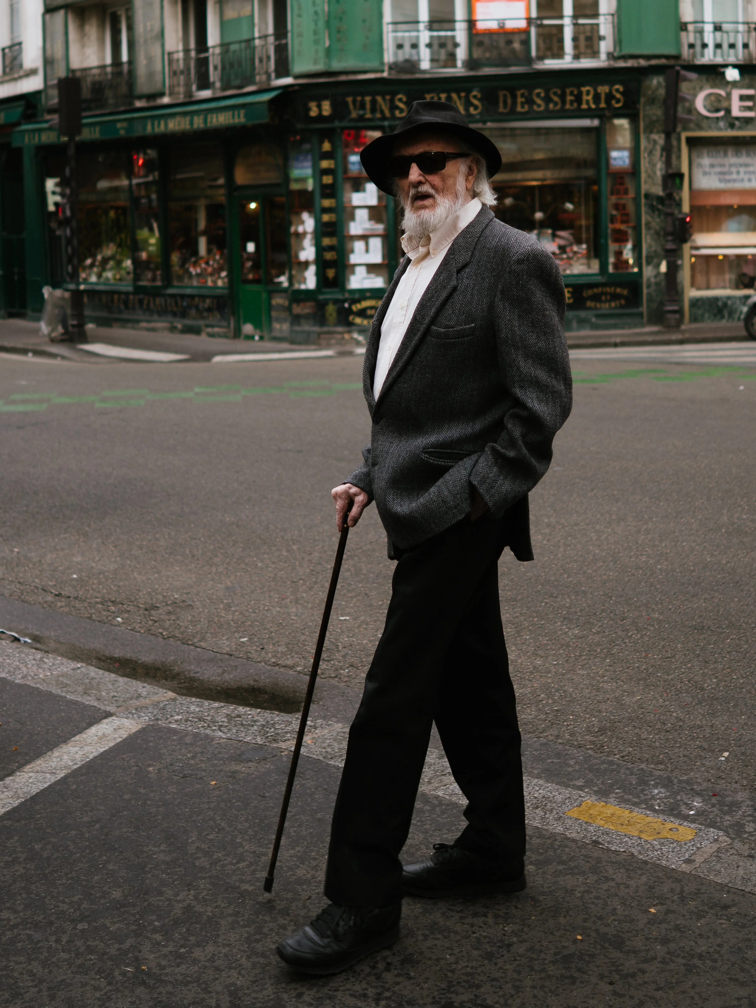 Elderly man with white hair and beard walking across a city street, dressed in a gray blazer, white shirt, black pants, and black shoes, holding a cane, wearing sunglasses and a black fedora hat, with a background of shops including a wine and desser