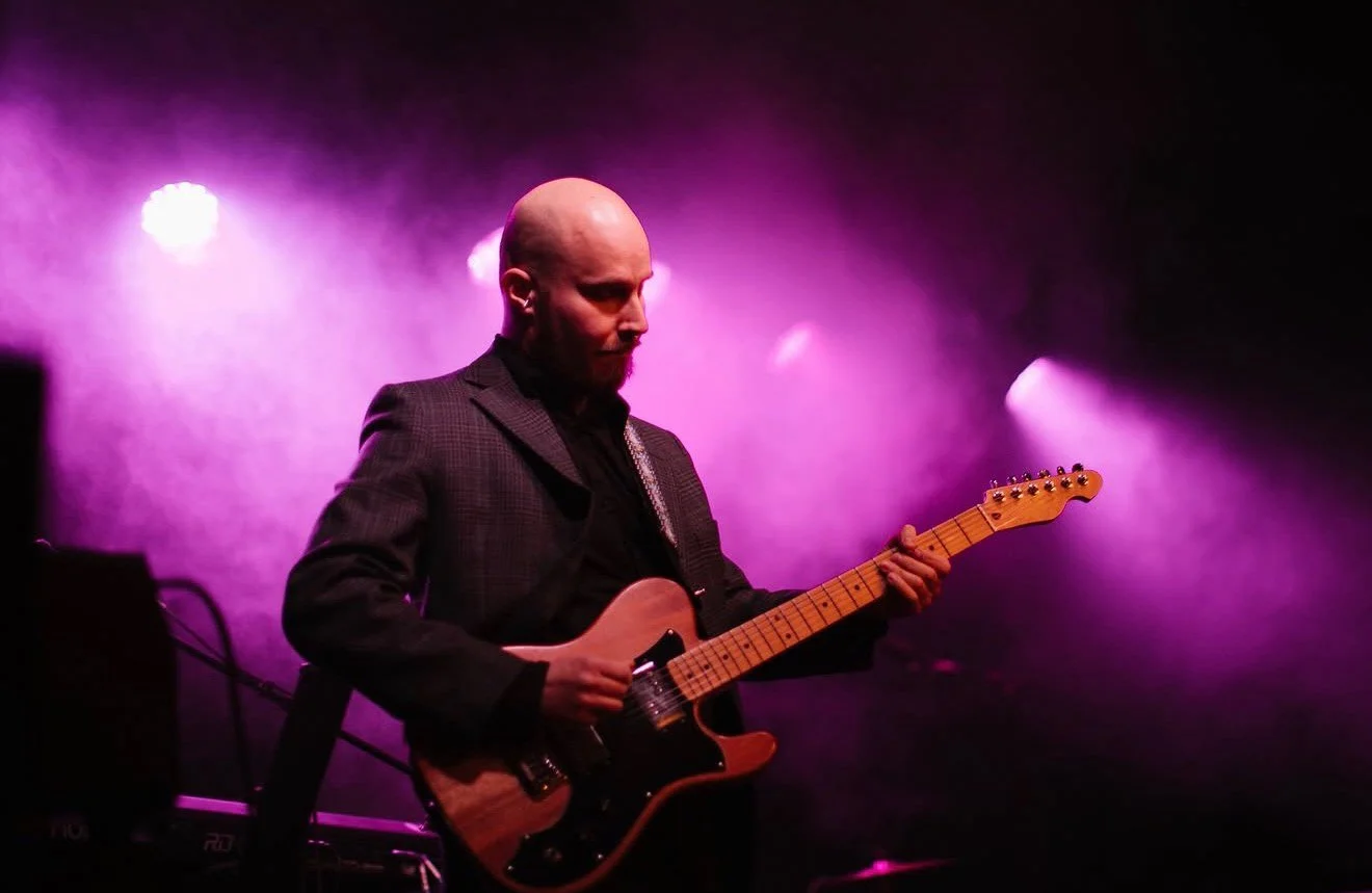 A man playing an electric guitar on stage with purple stage lighting and smoke effects.