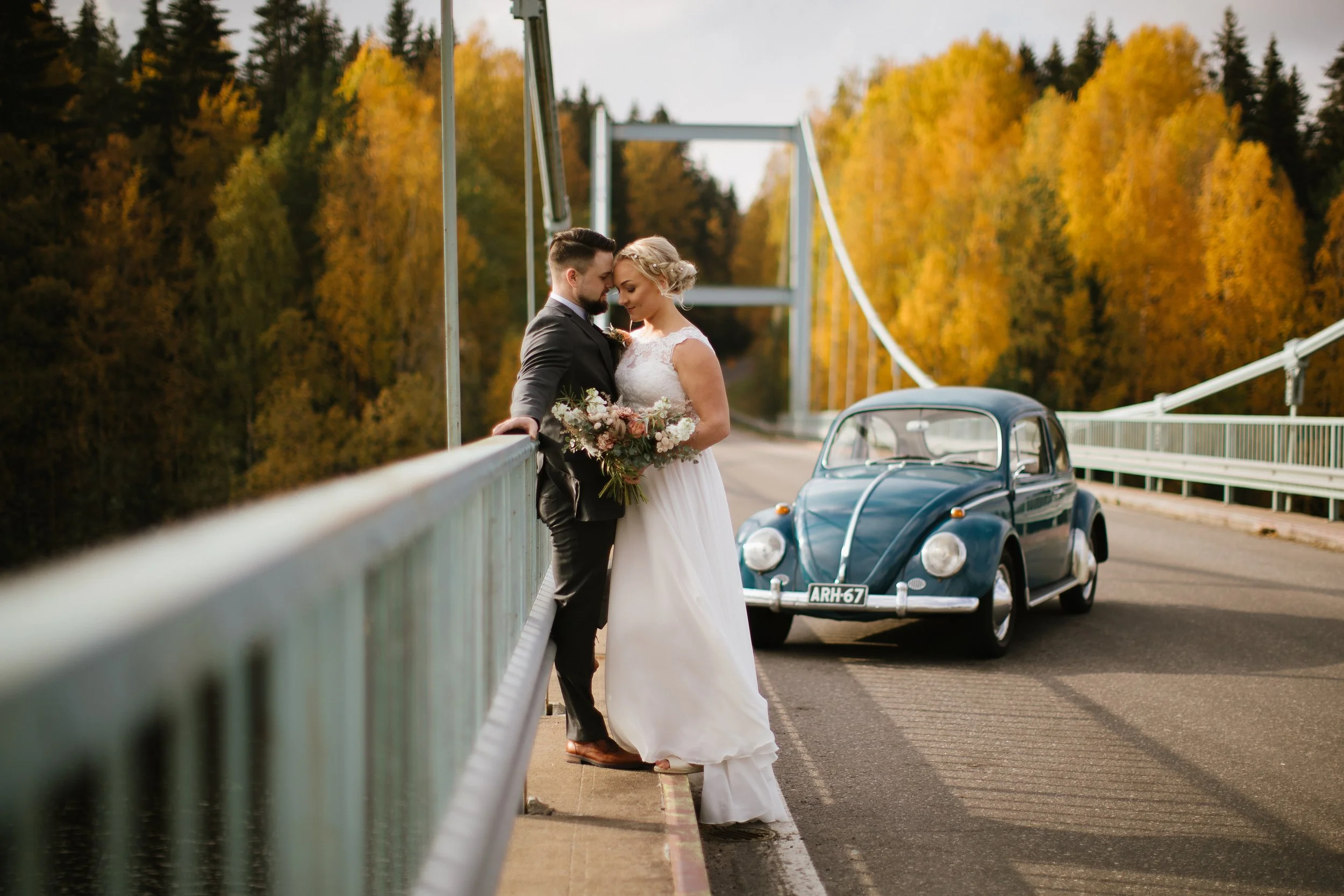 A bride and groom leaning their foreheads together on a bridge during a wedding photoshoot, with a vintage blue car parked nearby and a backdrop of autumn-colored trees.