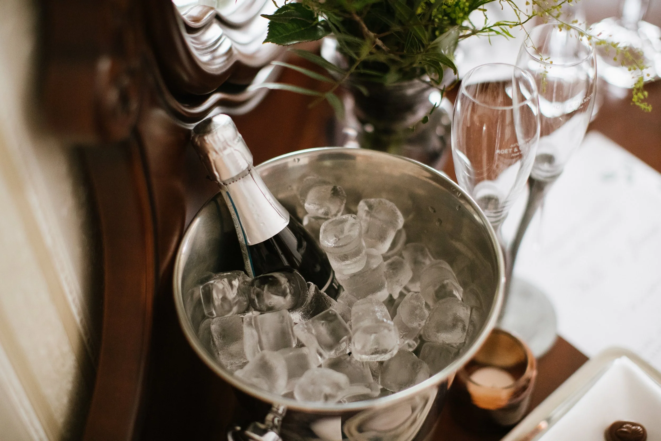 Wine bottles in an ice bucket on a wooden table with a vase of green foliage and flowers nearby.