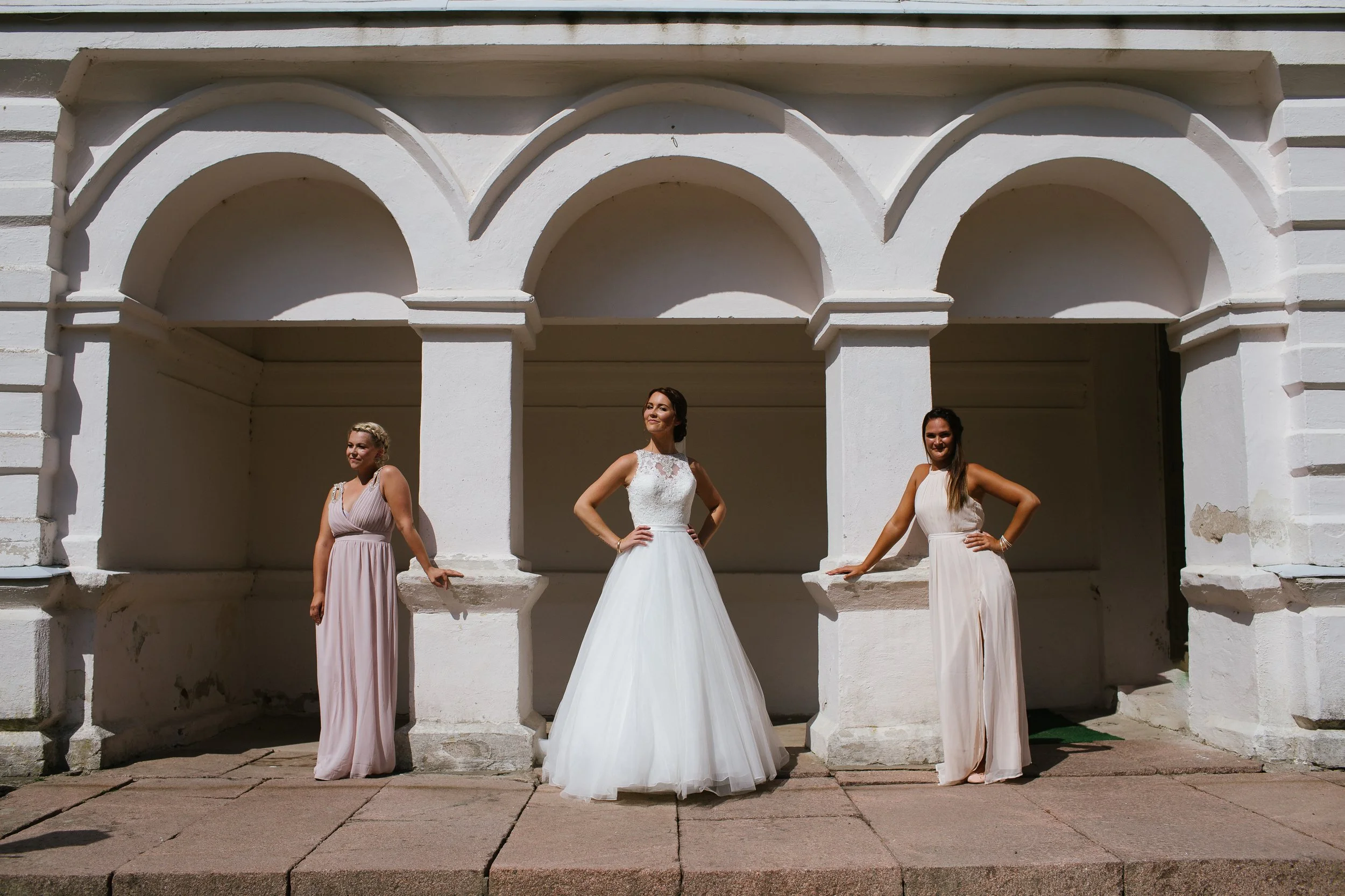 Three women in formal dresses standing in front of a white stone arch structure during daytime.