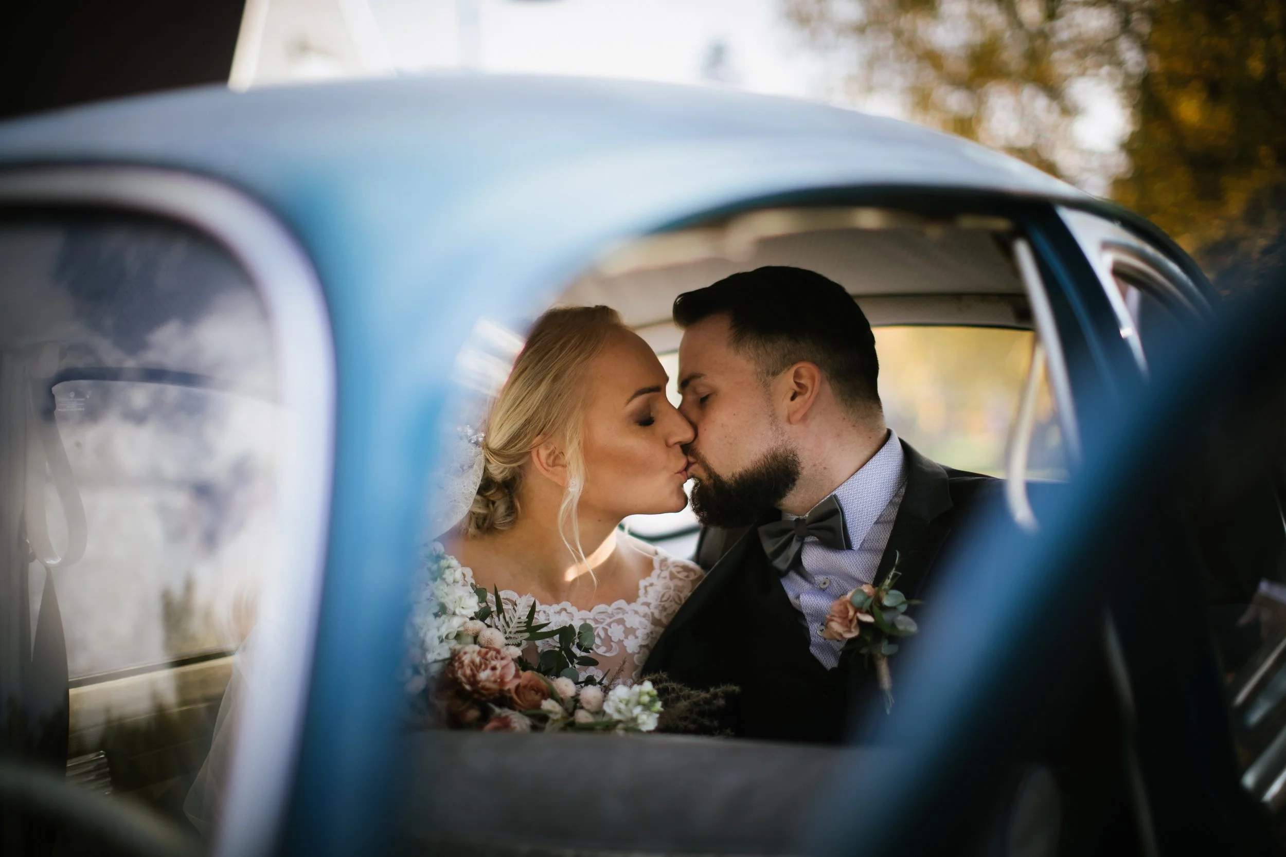 A bride and groom share a kiss inside a vintage car, seen through the window with blurred autumn trees in the background, during their wedding.