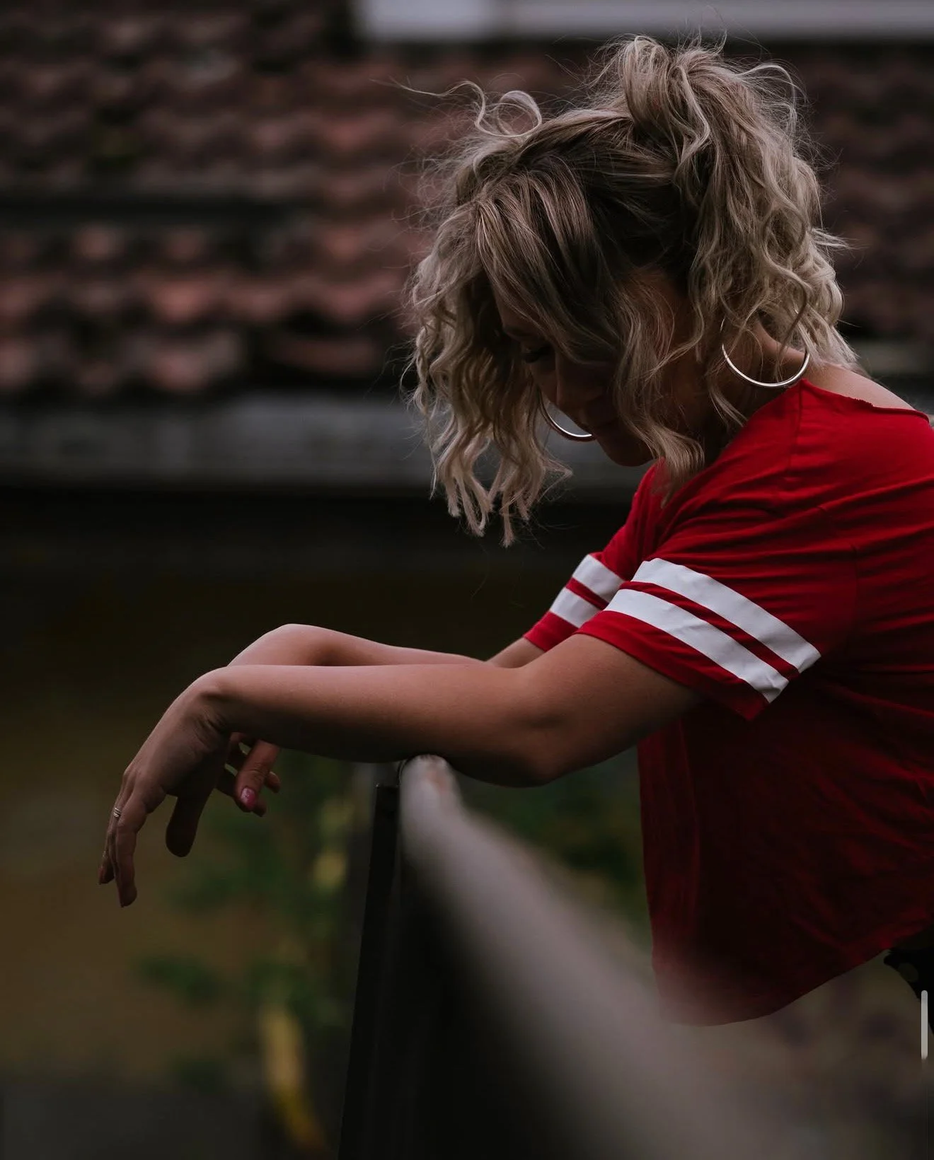 A woman with curly blonde hair wearing a red shirt with white stripes on the sleeves, leaning on a railing outdoors with a blurred background.