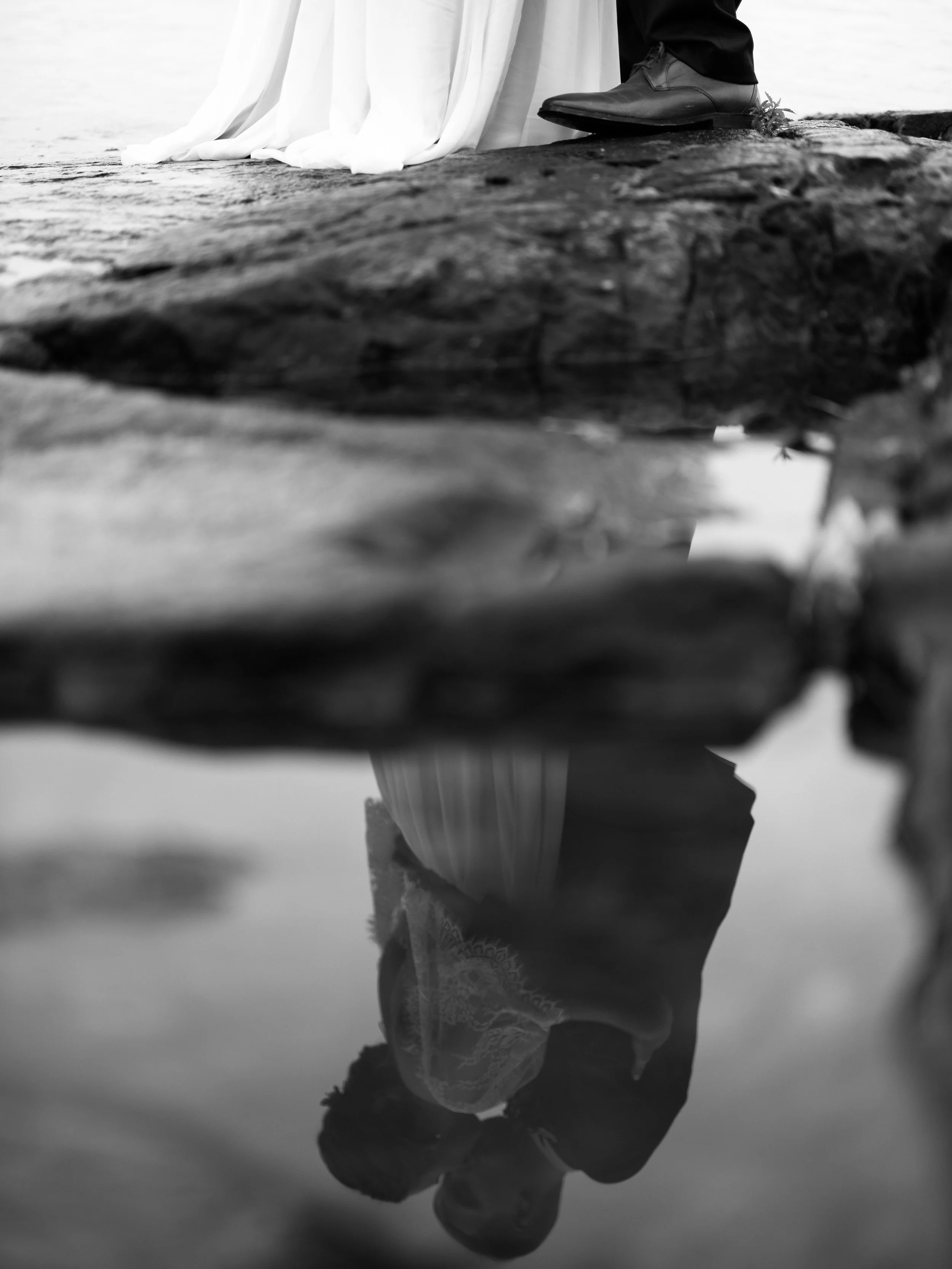 A black and white photo of a person's reflection in a puddle, showing their face and upper body, with the person wearing dark clothing and standing on a textured surface.