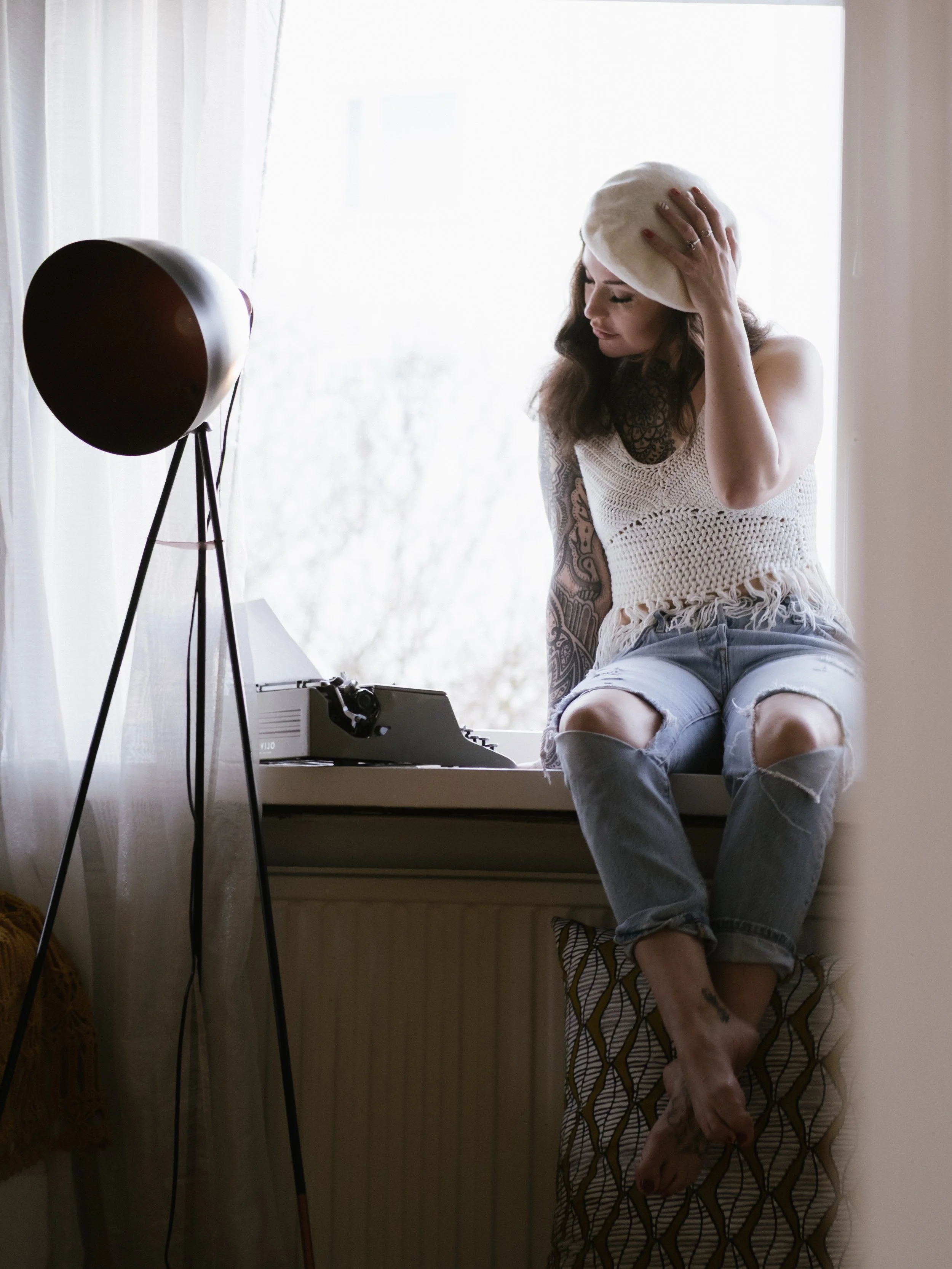 A woman with long dark hair, tattoos, and a beige beret sits on a window seat in front of a window with white curtains. She is wearing a crocheted white top and ripped jeans, holding her head with one hand.