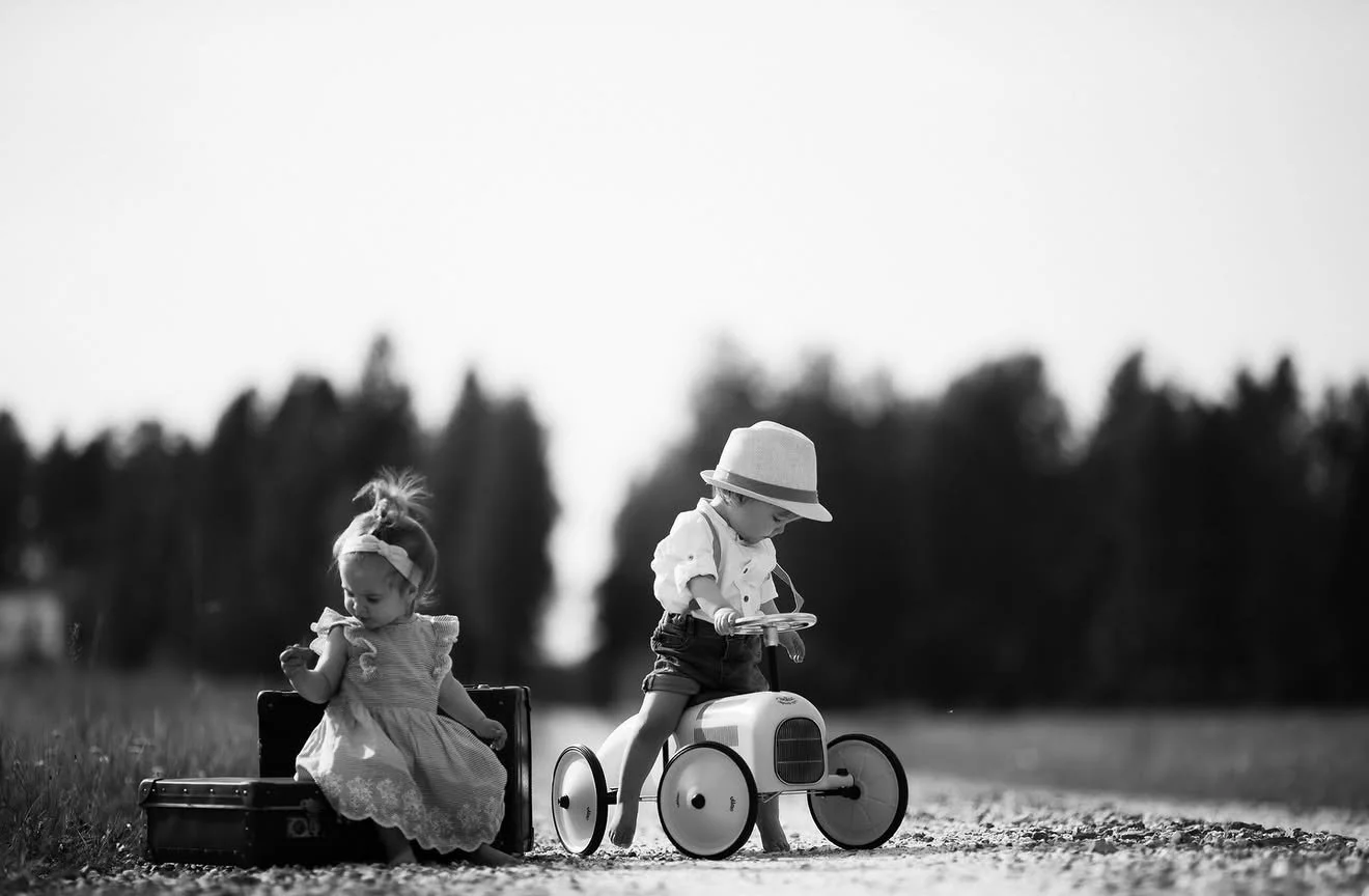 Black and white photo of two young children playing outdoors on a gravel path. One girl is sitting next to an open suitcase, looking down, and the other boy is riding a small vintage wheelchair. The background is blurred with trees, and the scene evo