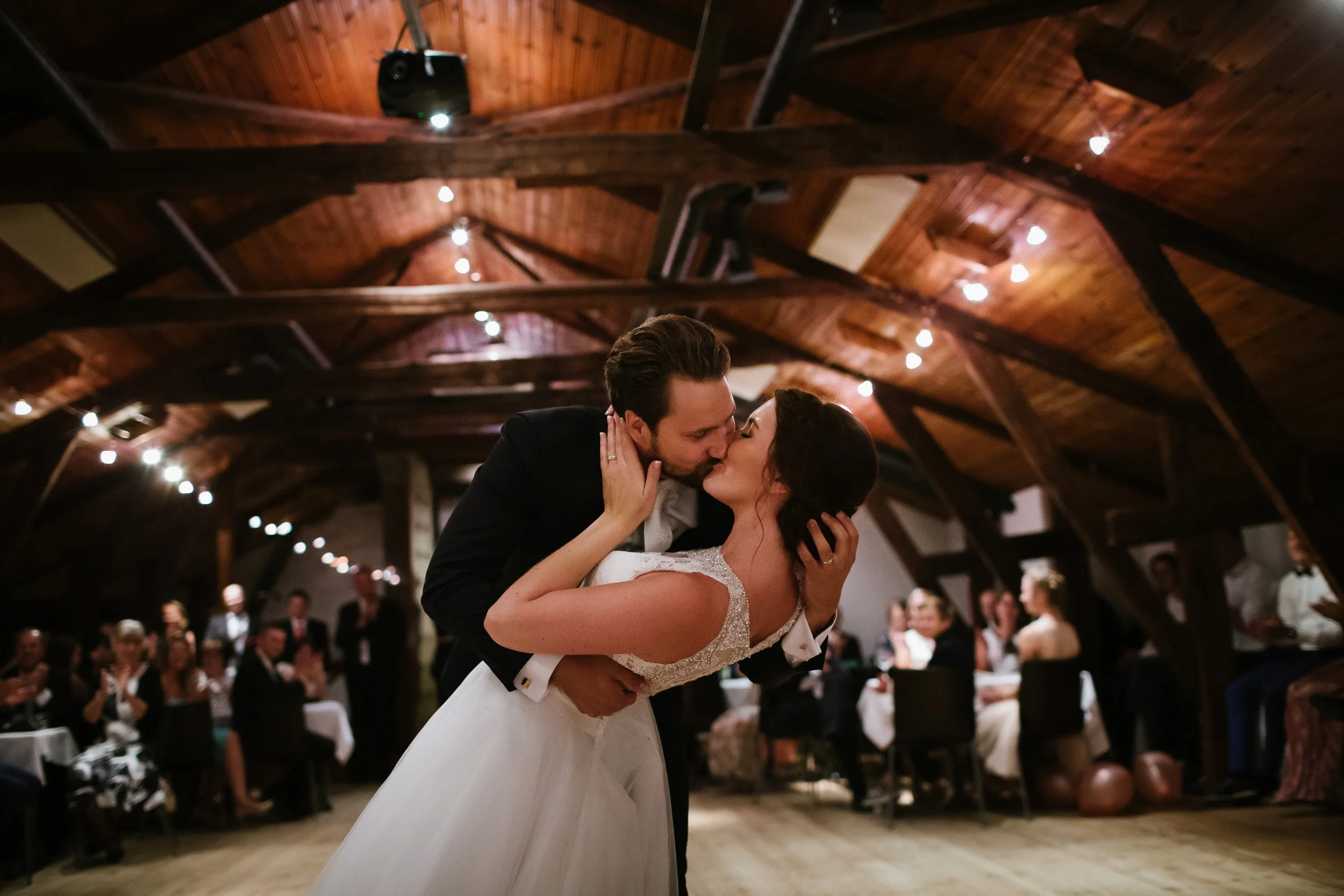A bride and groom share a dance during their wedding reception in a rustic wooden venue with wedding guests watching.