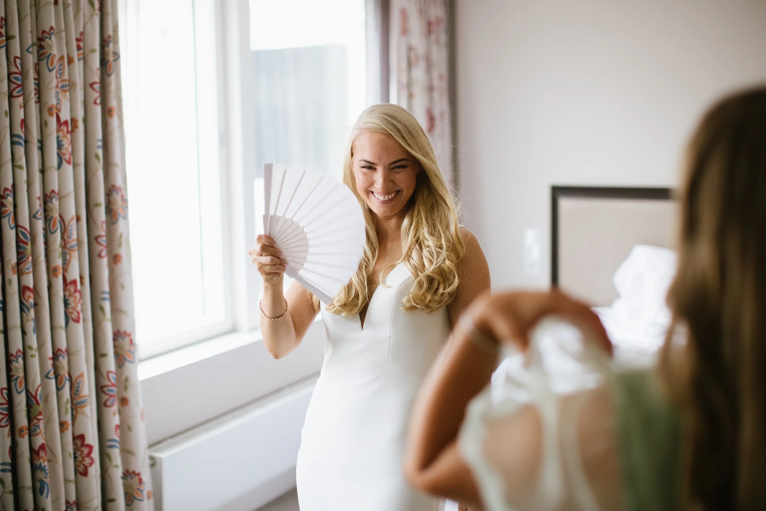 A woman with long blonde hair smiling and holding a white fan inside a well-lit room, with curtains and a bed in the background.