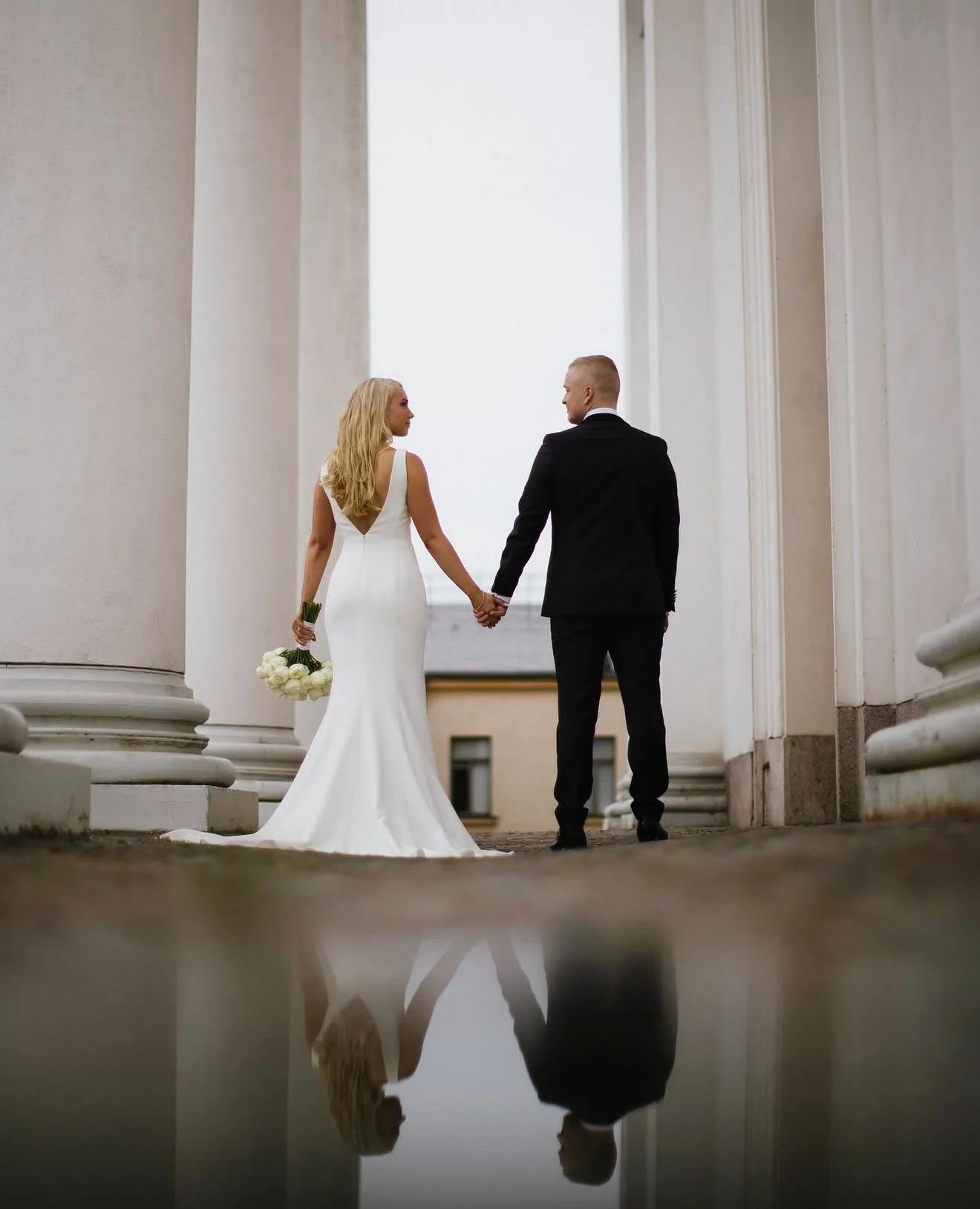 A bride and groom walk hand in hand between large white columns, with a reflection of them on a shiny surface below.