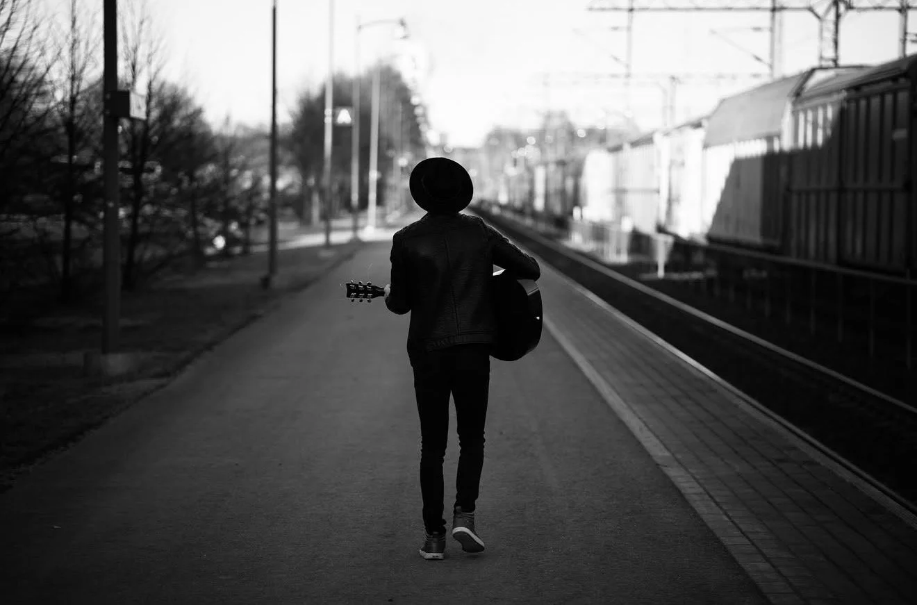 A person walks along a train platform while holding a guitar, wearing a hat and jacket, with train cars in the background.