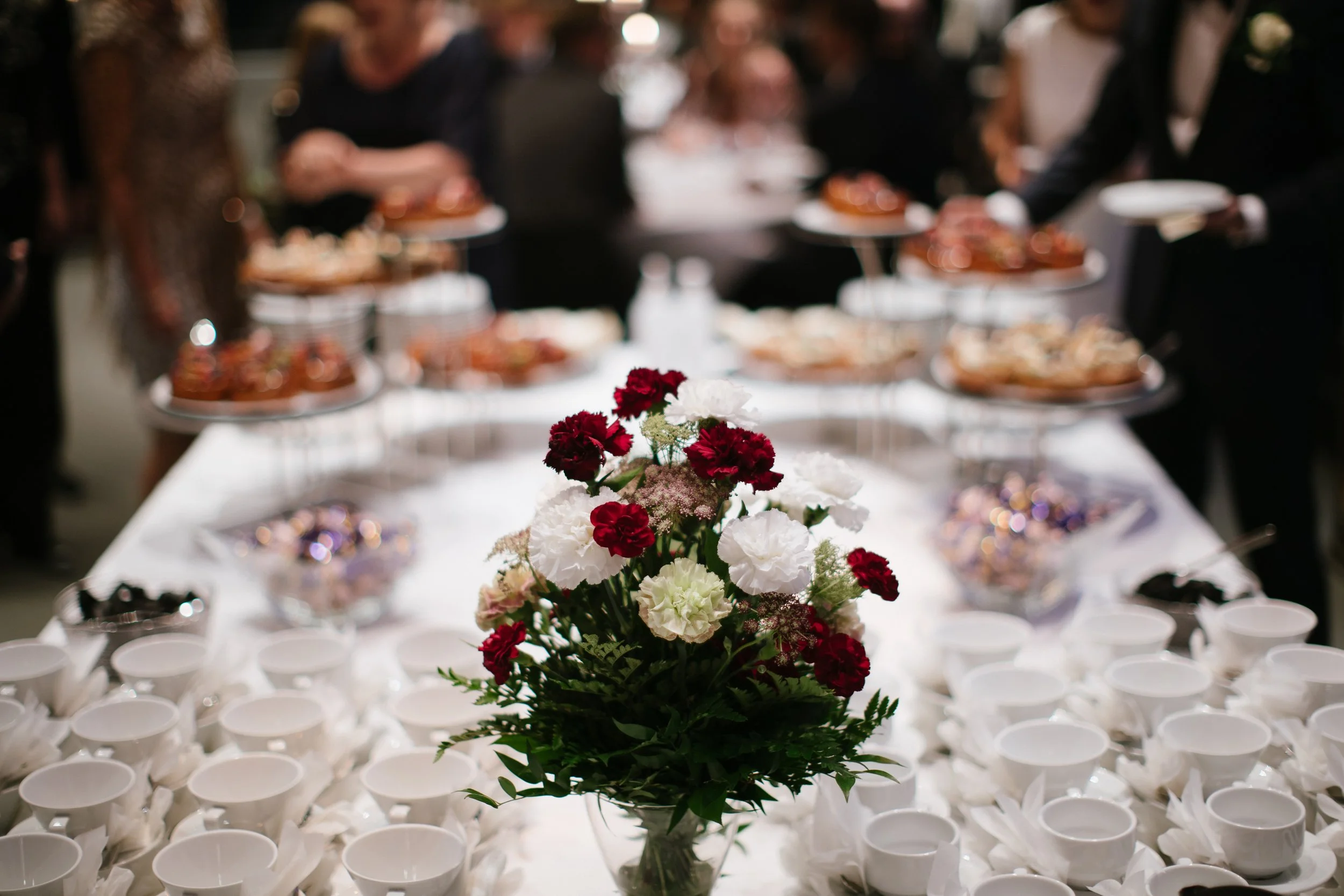 A flower arrangement with red and white carnations on a table with numerous empty white cups, blurred people in formal attire in the background at a social event.