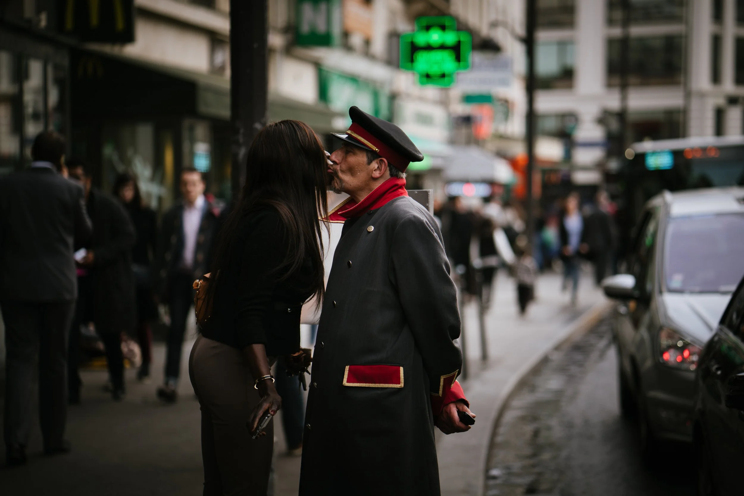 A man dressed as a bellhop is kissing a woman on the lips on a city street, with people walking by and green cross pharmacy signs in the background.