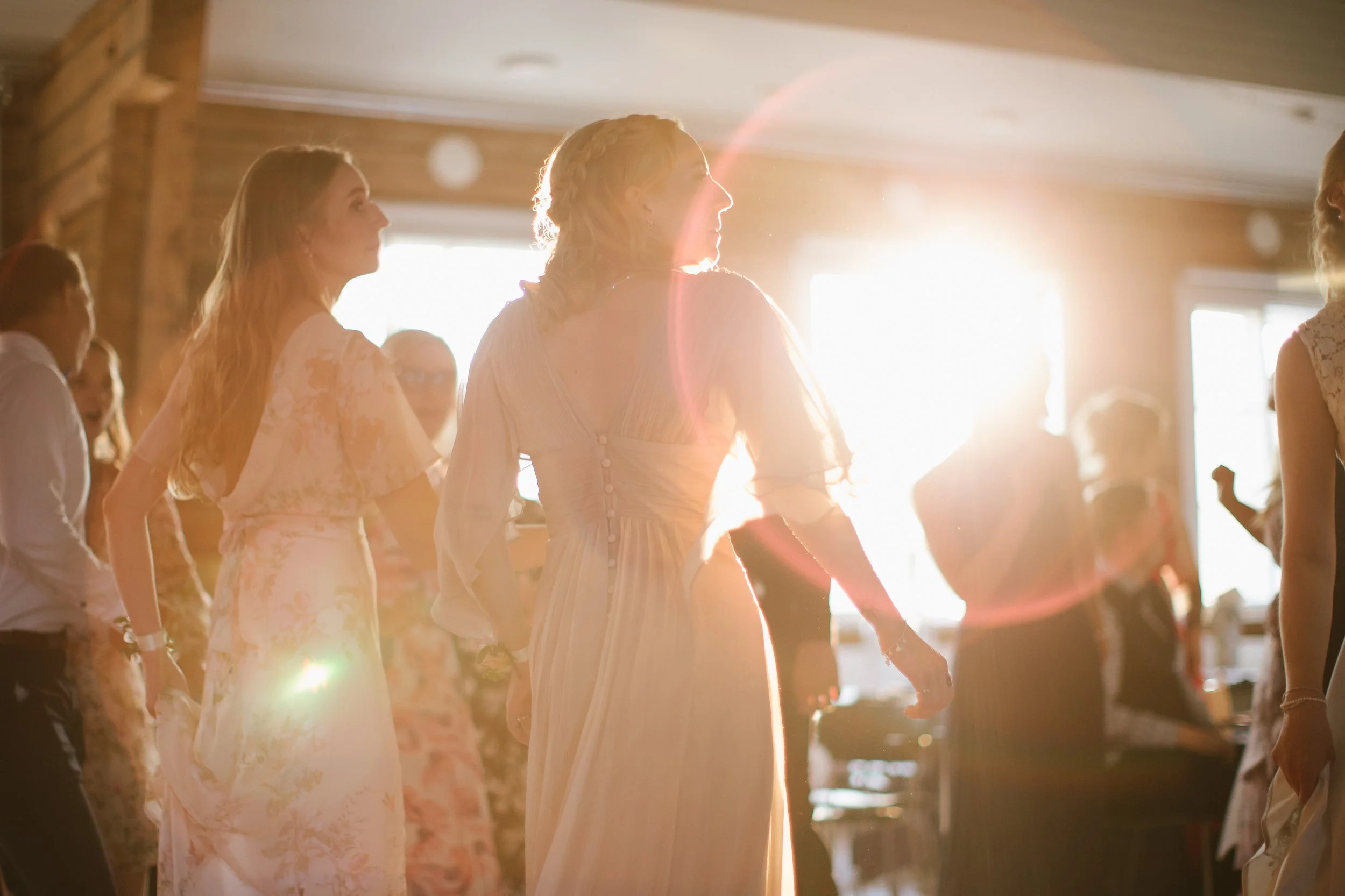 Group of women holding hands during a dance at a wedding reception, illuminated by bright sunlight streaming through windows.