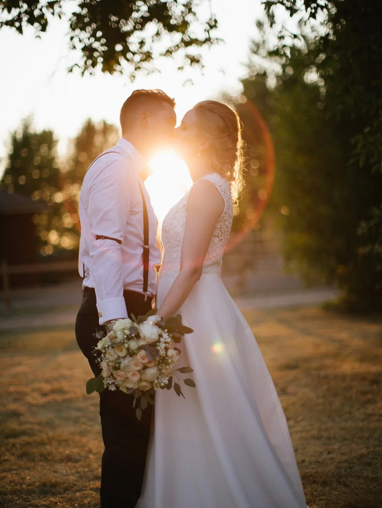 A bride and groom share a kiss outdoors at sunset, with the sun shining between them. The bride holds a bouquet of white and blush flowers, and they are dressed in wedding attire. The scene is romantic and serene with trees in the background.
