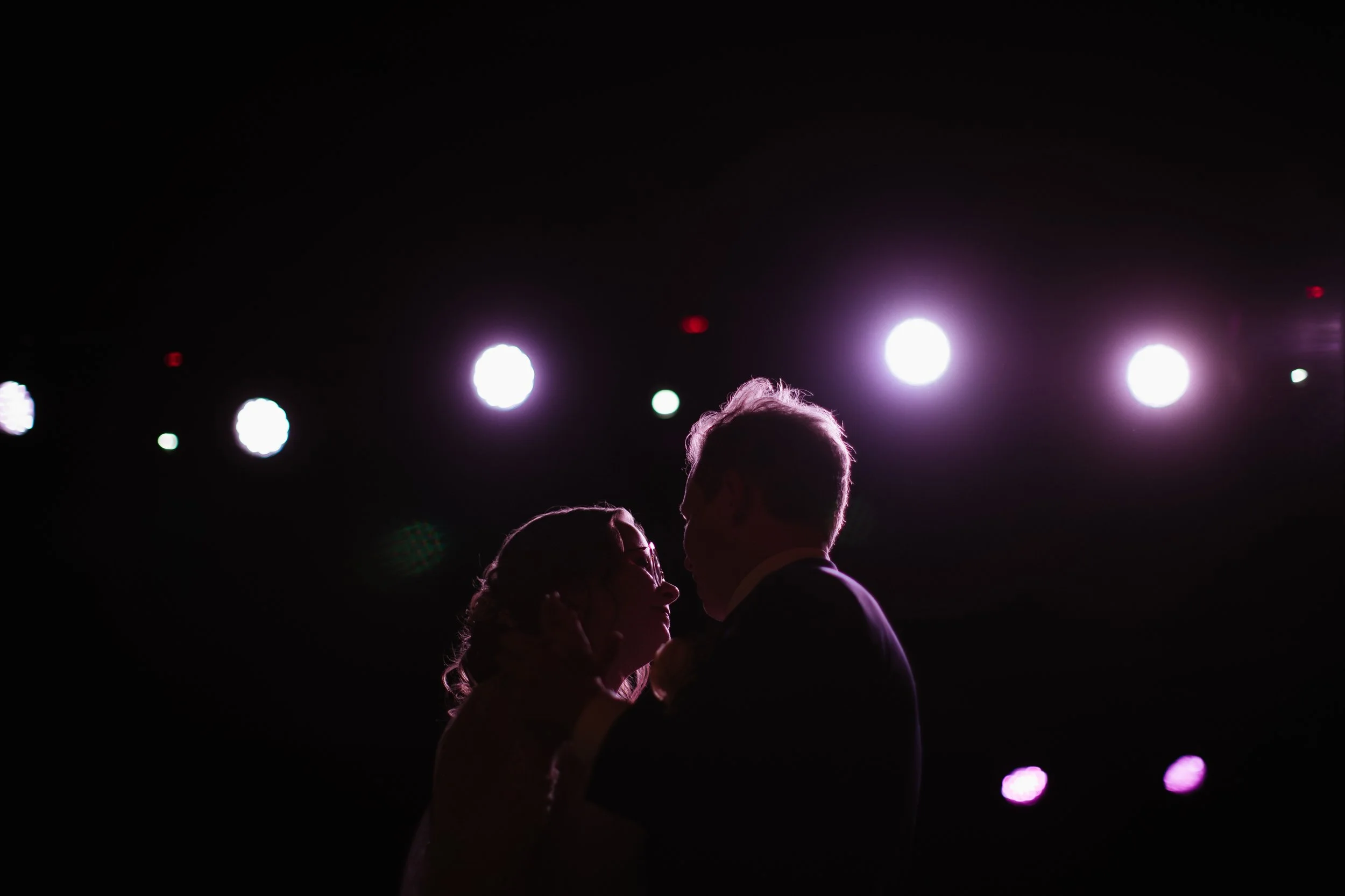 Silhouette of a couple dancing closely at an event with bright stage lights in the background.