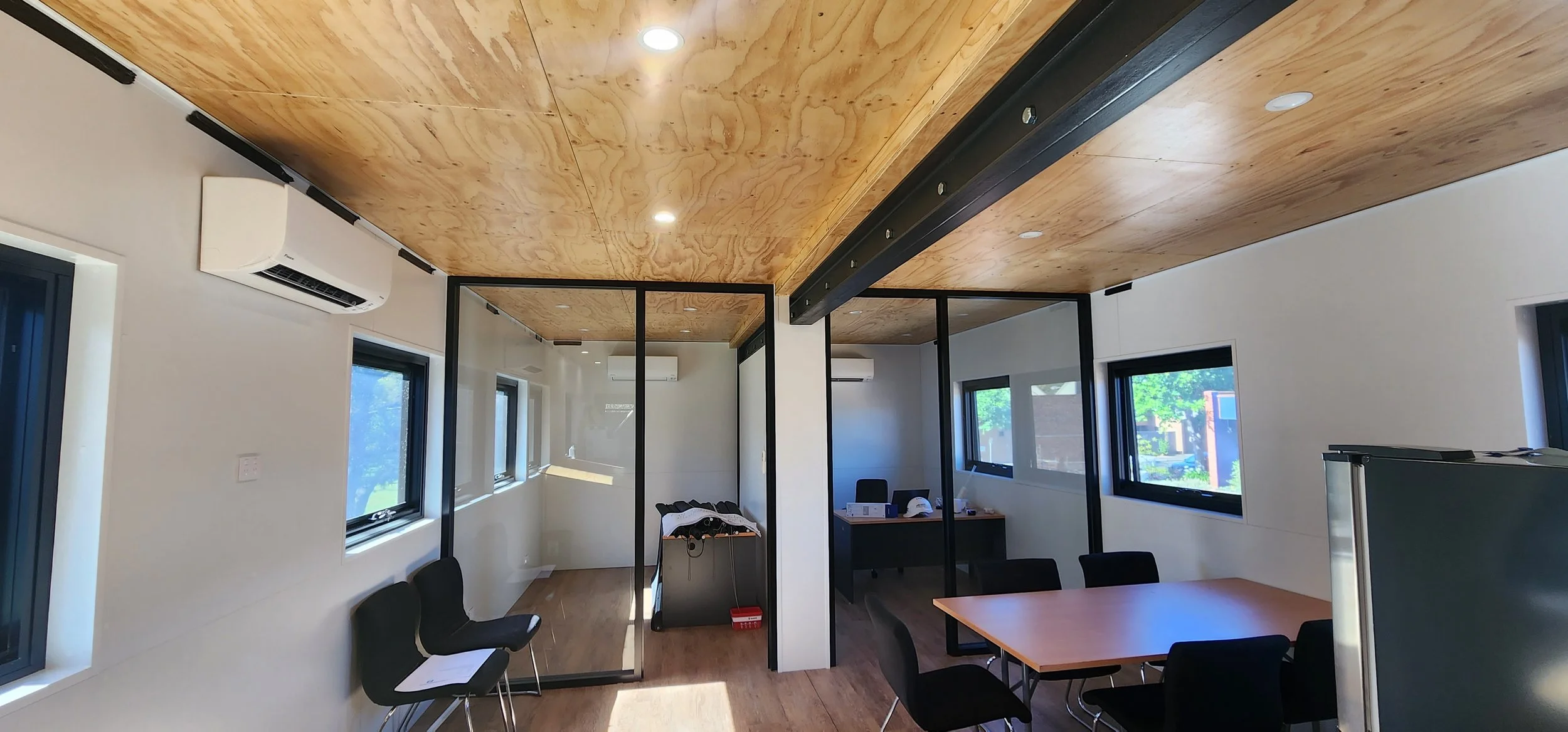 Modern office interior with wooden ceiling, white walls, multiple windows, black chairs, and glass-partitioned rooms.