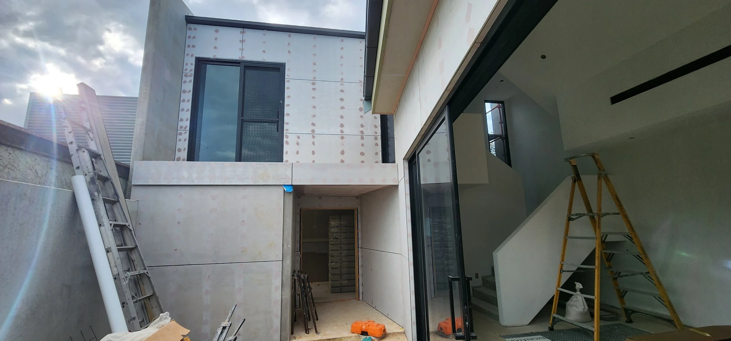 Construction site of a modern building with large glass windows, scaffolding, and tools inside. The sky is partly cloudy.