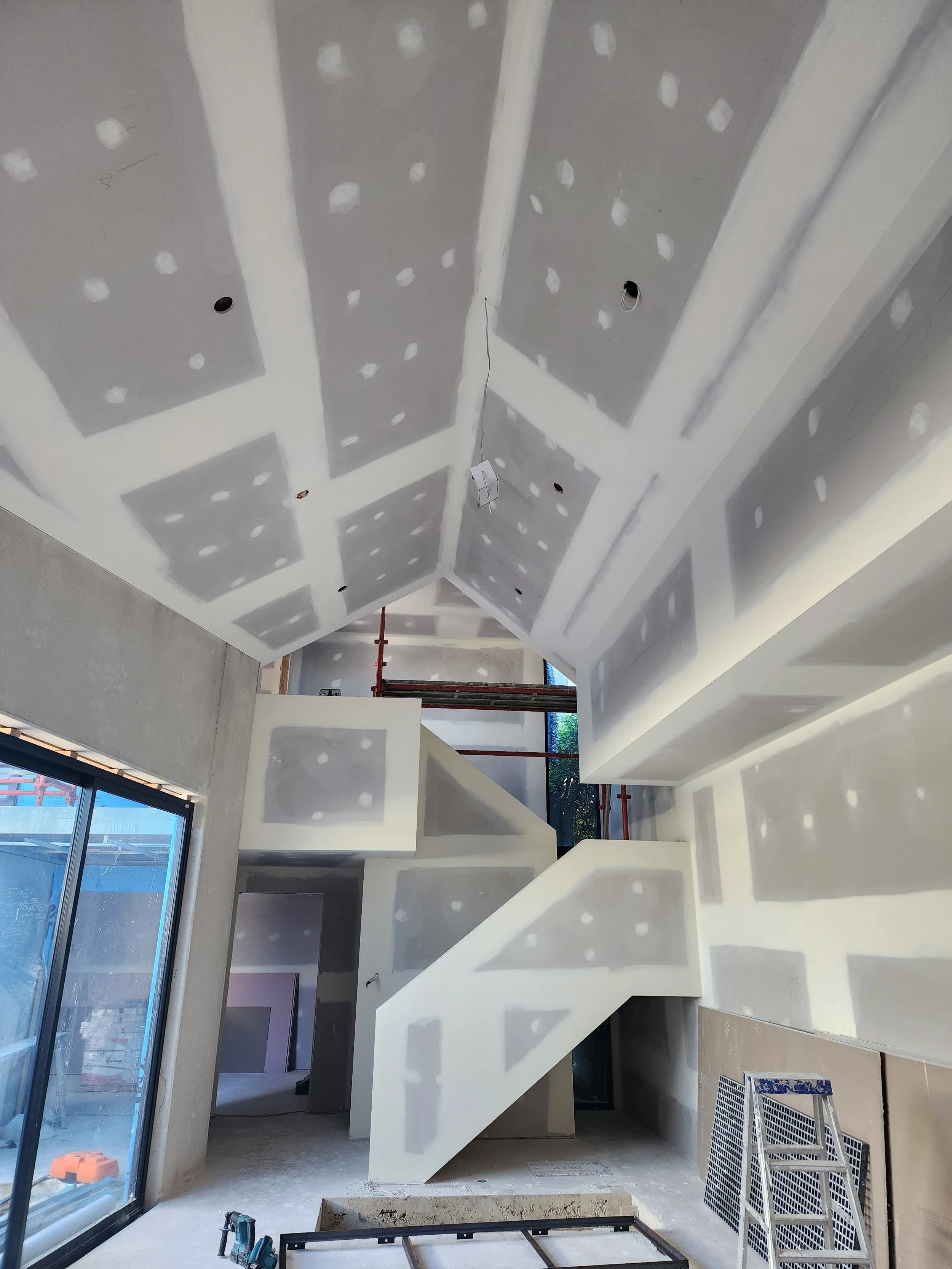 Interior view of a building under construction, showing drywall installation on the ceiling and walls, with some electrical wiring and tools present.
