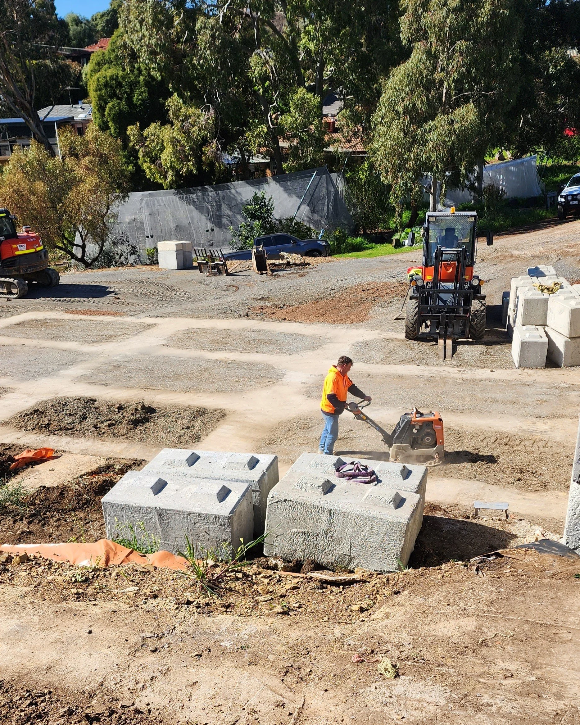 Construction site with a worker operating a compactor, large concrete blocks, trees, construction vehicles, and a partially developed dirt road.