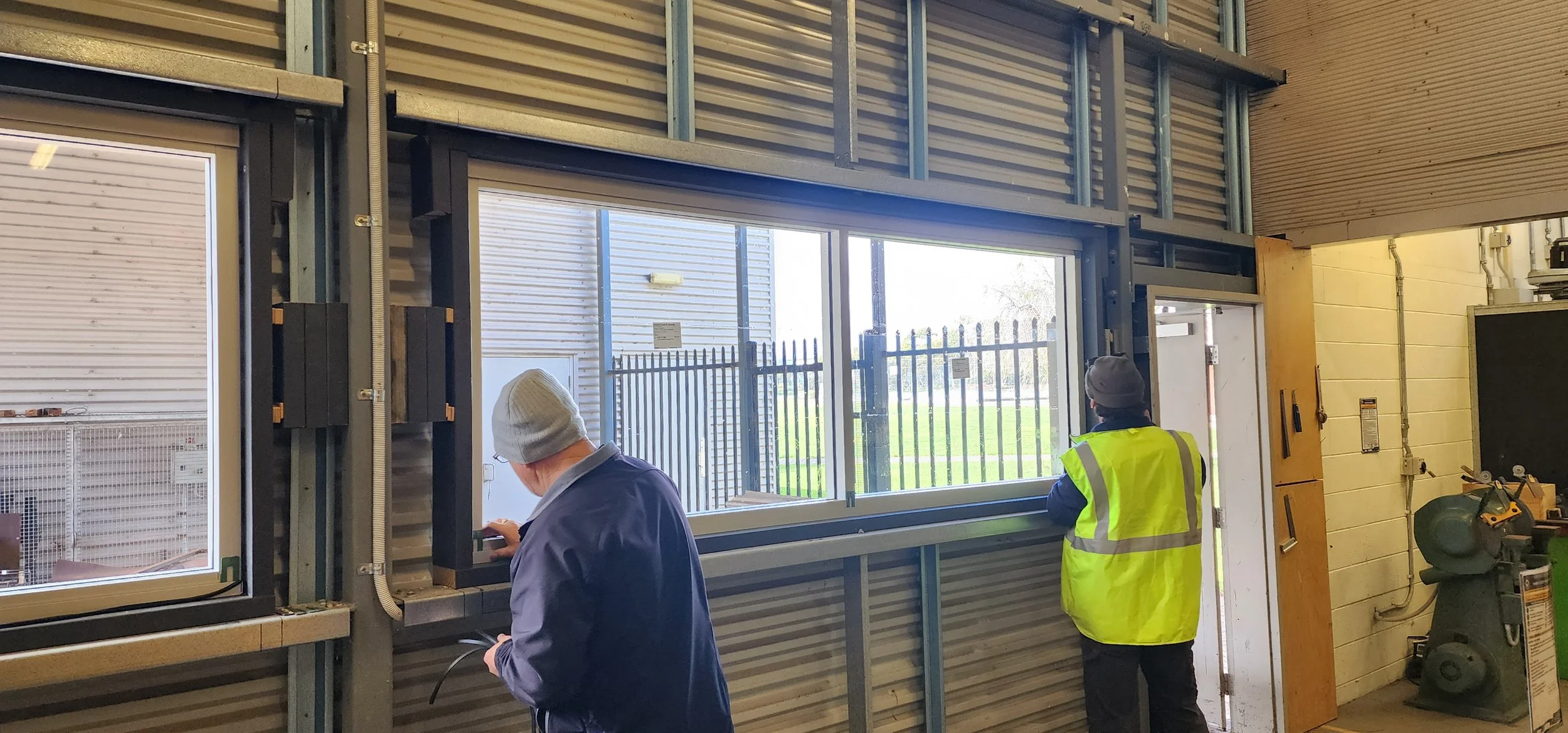 Two workers installing or repairing a large window in an industrial setting. One worker is wearing a gray beanie and navy jacket, working on the lower part of the window. The other is wearing a gray beanie and a yellow safety vest, facing away from the camera. The window shows an outdoor area with a grass yard and a metal fence.