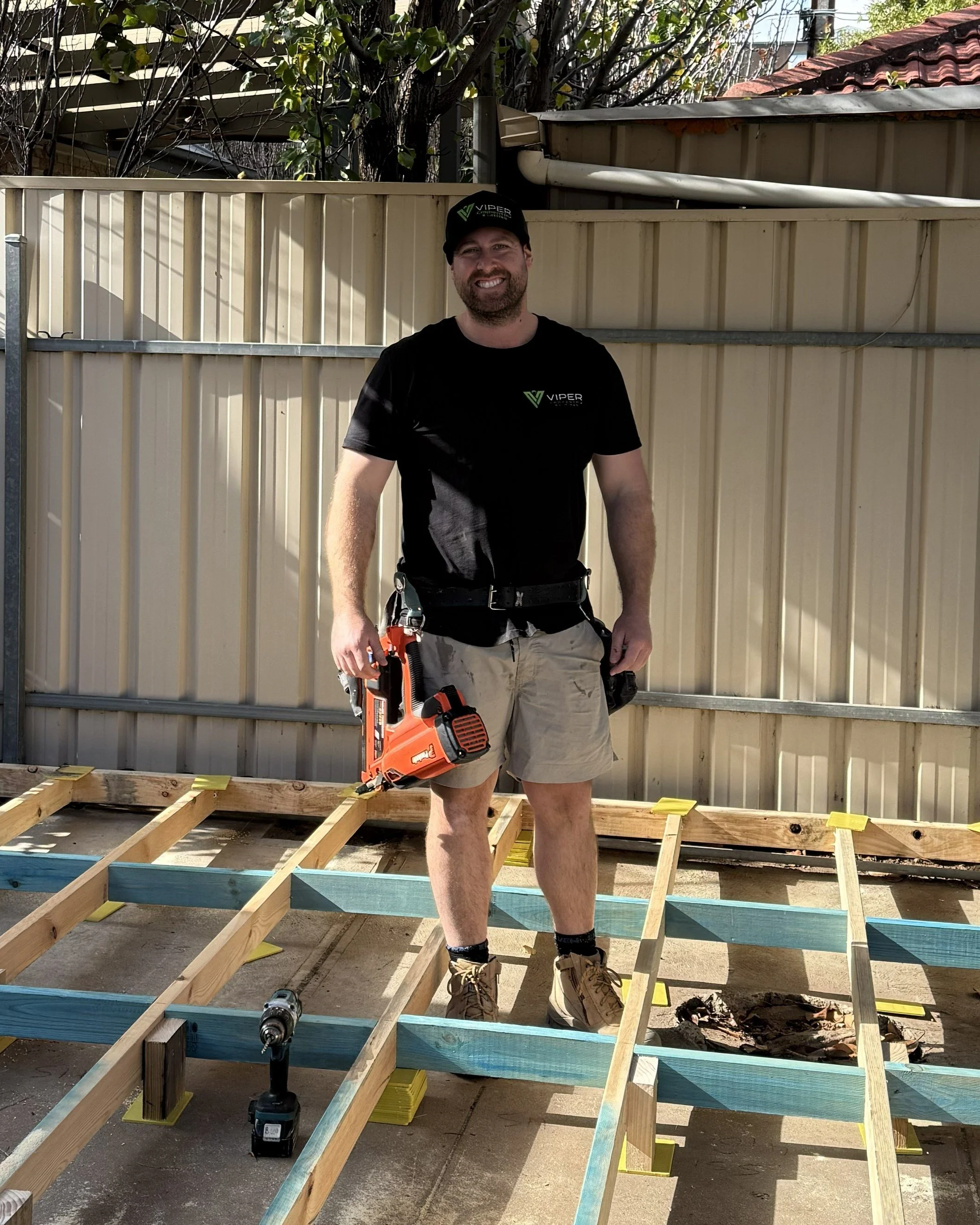 A man standing outdoors in front of a beige fence and wooden foundation frame, holding a power tool, wearing work boots, shorts, a black t-shirt and a black cap, during daytime.