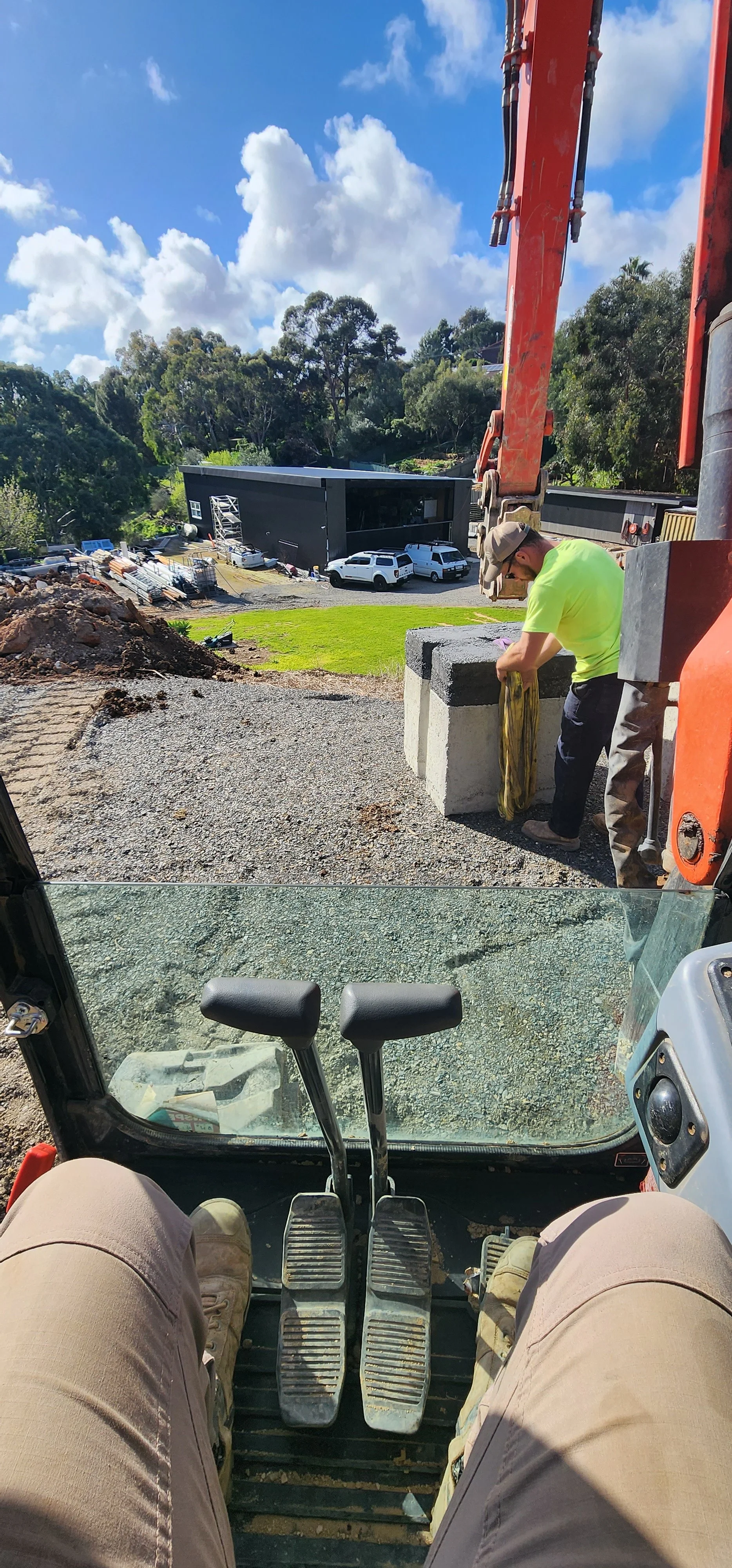 The photo shows the view from inside a construction vehicle, looking out over a construction site with a worker standing next to a concrete block, and a modern black building in the background. The person's legs and controls of the vehicle are visible in the foreground.