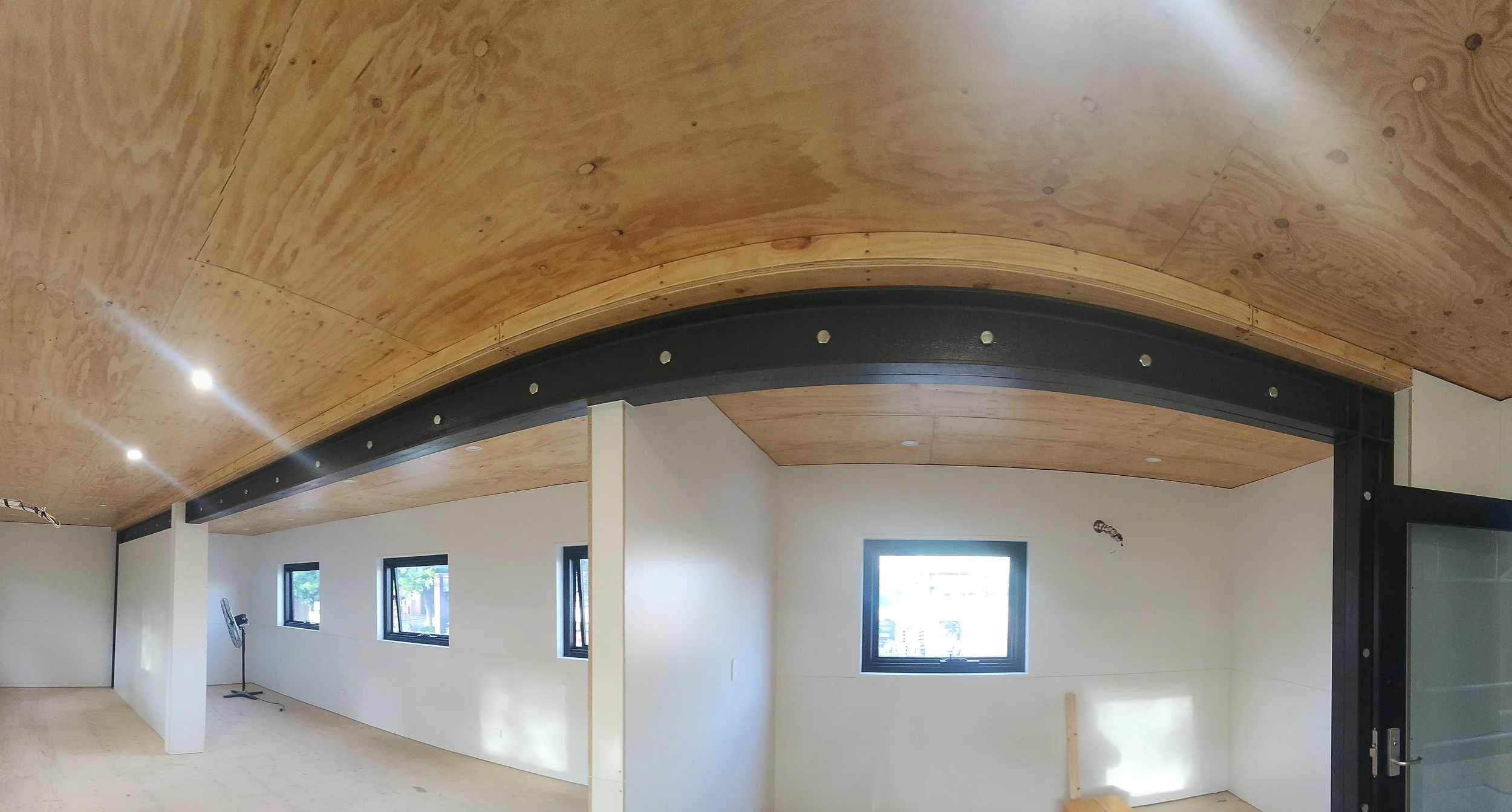 Interior view of a room with a curved wooden ceiling, black beams, multiple windows, and white walls.