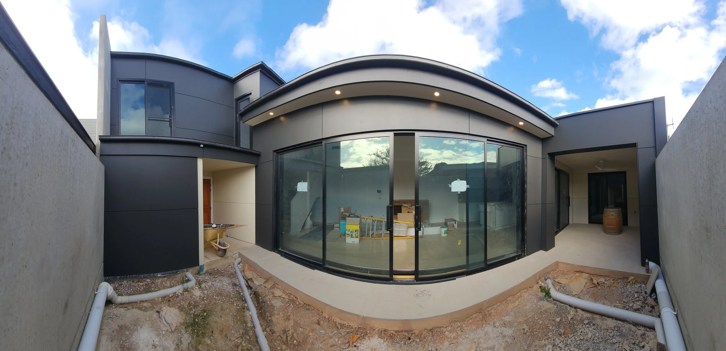 Modern house under construction with large glass windows, surrounded by concrete walls and construction materials.
