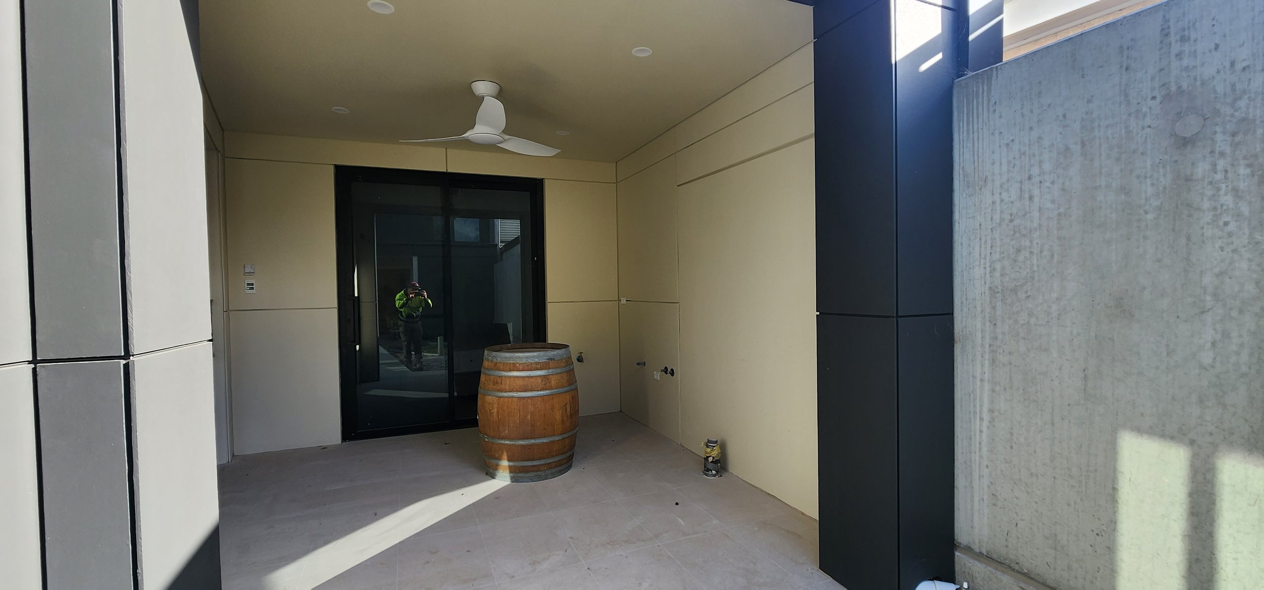 Empty covered patio space with a ceiling fan, a door with glass panels, a large barrel, and some construction materials on the floor.
