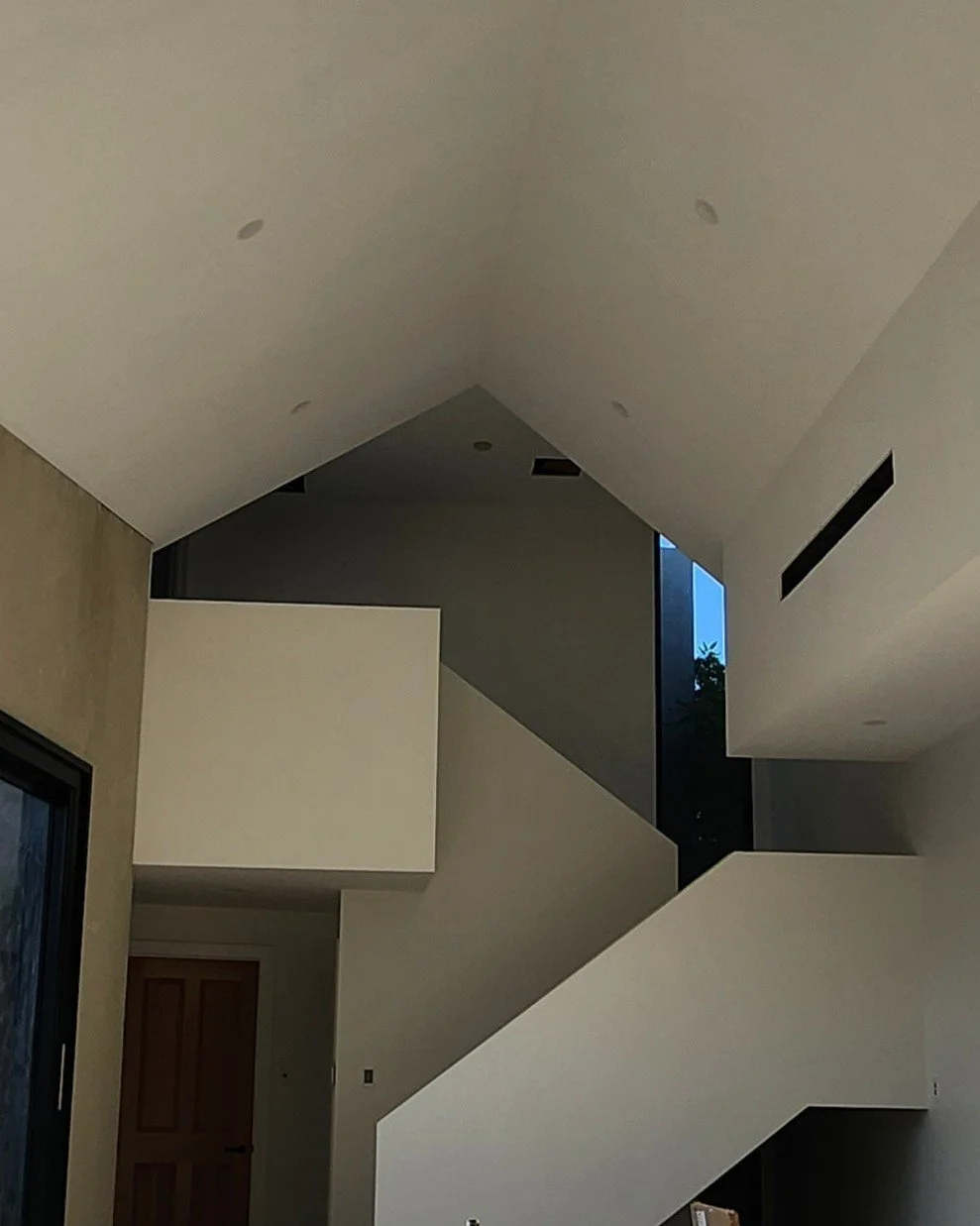 Interior view of a modern house with white walls, a high ceiling with recessed lighting, and large windows showing trees outside.