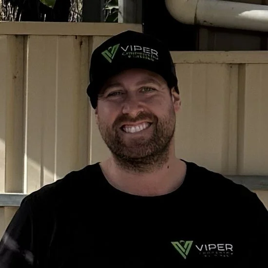 A smiling man wearing a black Viper Cap and T-shirt, standing outdoors in front of a wooden fence.