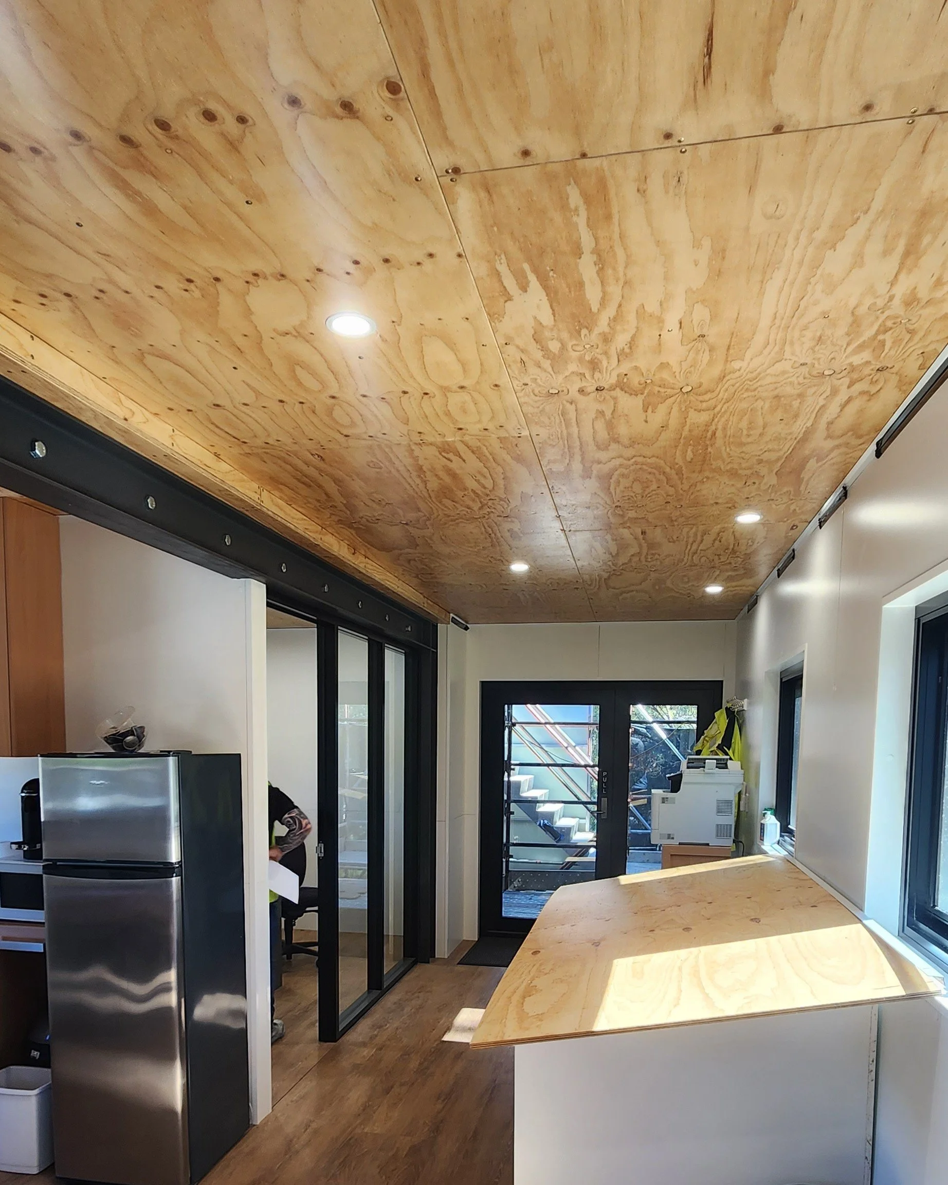 Interior view of a modern kitchen and dining area with a wooden ceiling, black sliding glass doors leading outside, a stainless steel refrigerator, a wooden dining table, and a view of outdoor stairs through the doors.