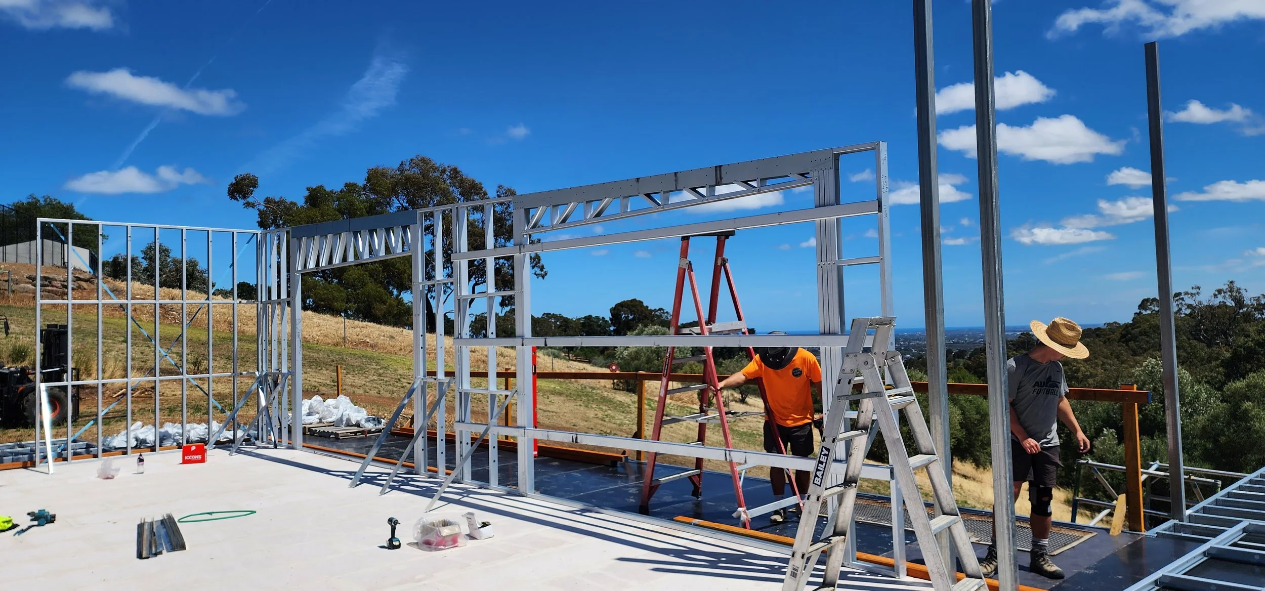 Two construction workers building a steel structure on a clear, sunny day with a blue sky and some clouds. One worker is wearing an orange shirt and black hat, the other is in a gray shirt and straw hat. Various tools and materials are scattered around the site.