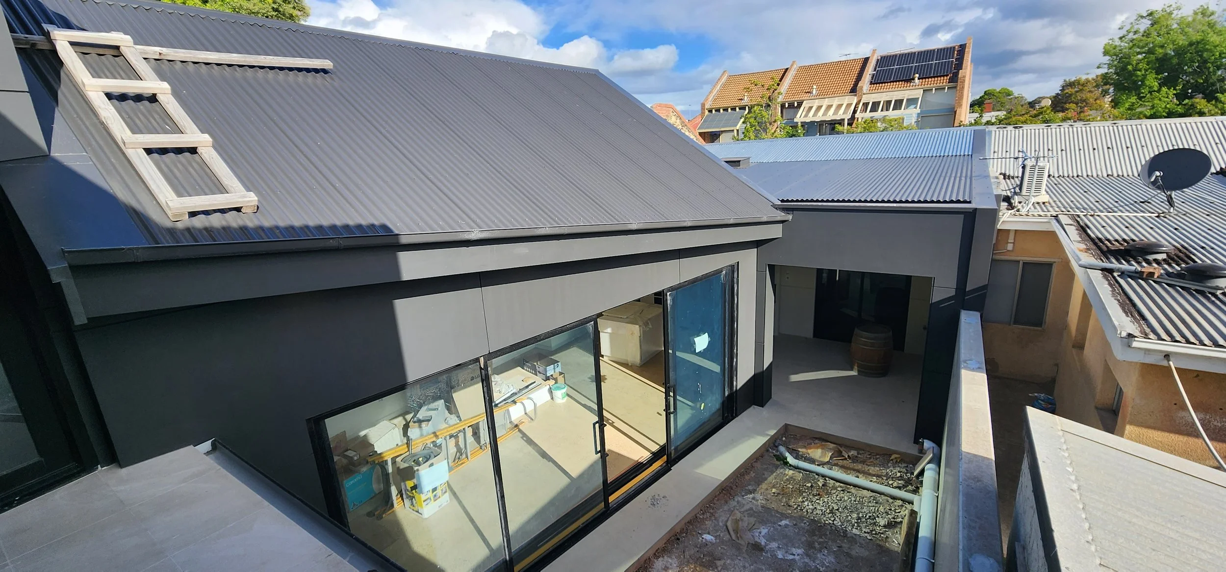 View of multiple rooftops with metal roofing, including a house under construction with sliding glass doors and some construction materials inside, during daytime with partly cloudy sky.
