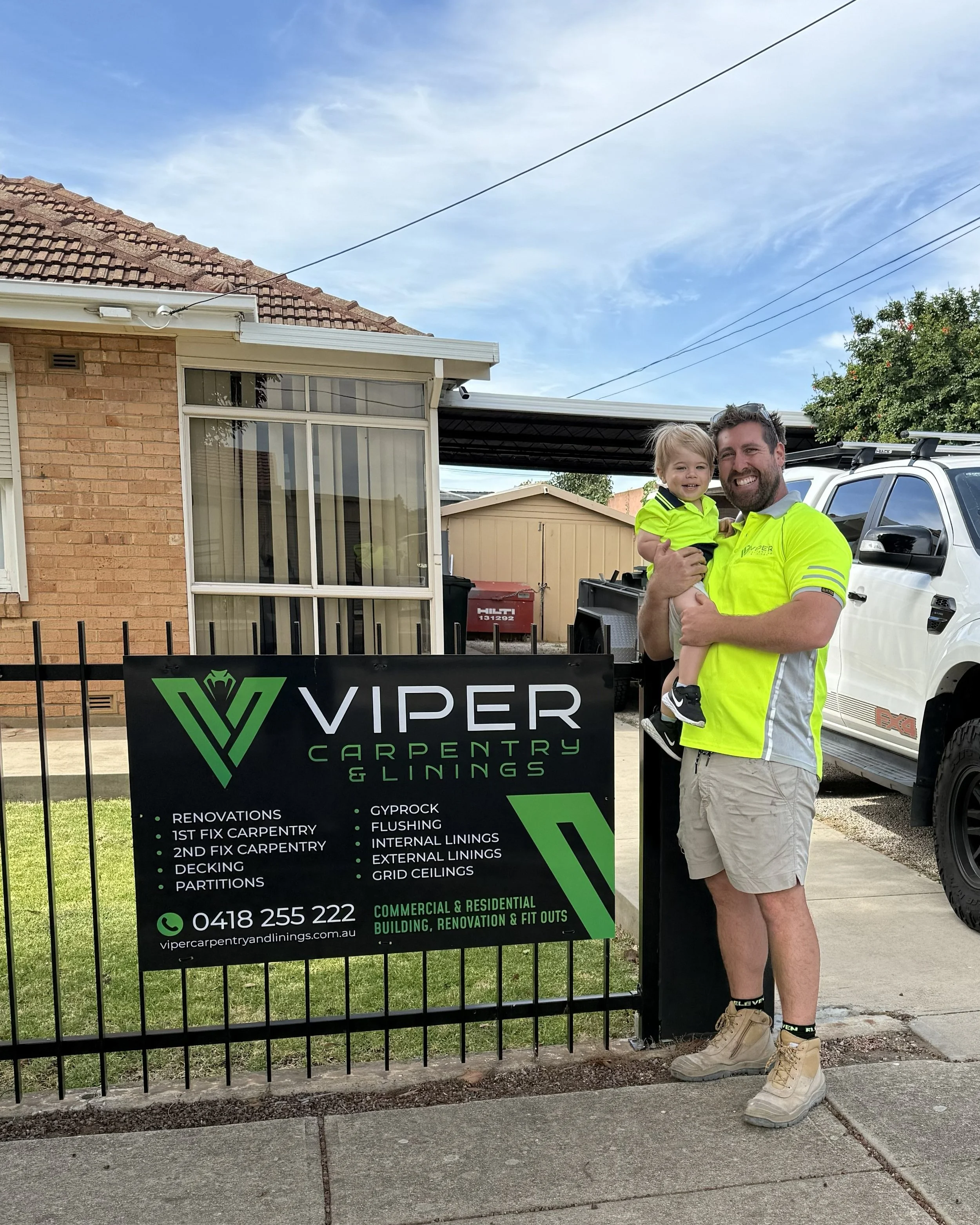 A man holding a smiling young boy in front of a sign for Viper Carpentry & Linings. The man is wearing a yellow high-visibility shirt and beige shorts, and the boy is wearing a neon yellow shirt. There is a white vehicle and a backyard with a house, a shed, and a tree behind them. The sign displays services offered by the company, including renovations, fixes, and linings.
