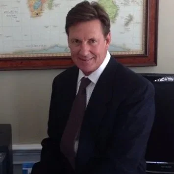 A man in a dark suit and tie sitting in an office with a world map on the wall behind him.
