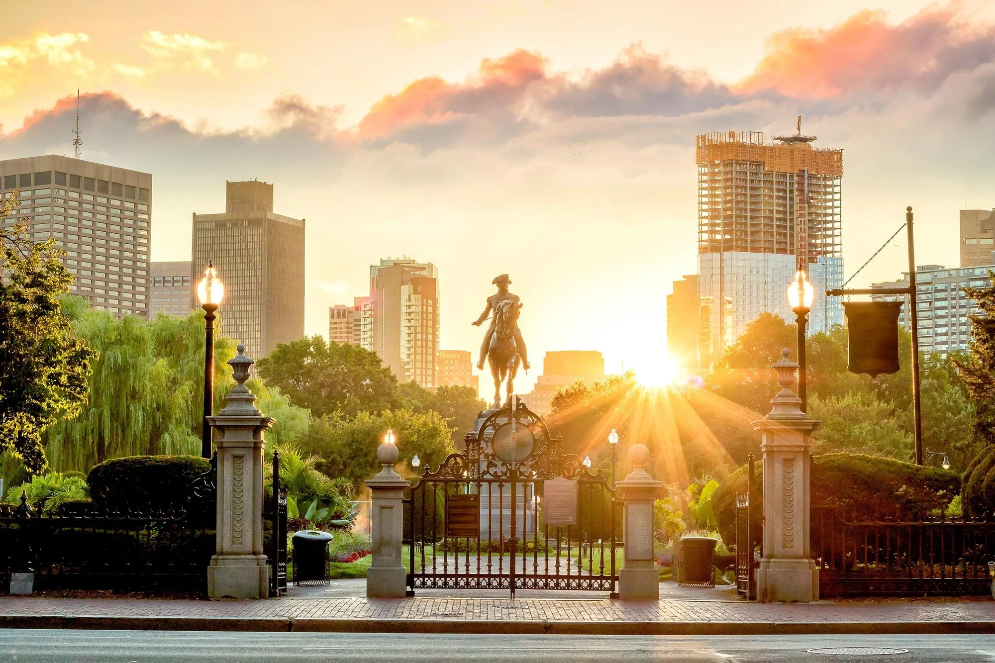 Sunset behind a city skyline with tall buildings, a statue on horseback in a park, decorative street lamps, lush greenery, and a gated entrance in the foreground.