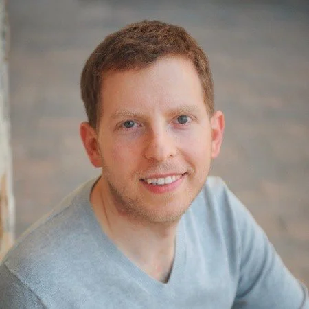 A smiling young man with short brown hair wearing a light gray shirt, posing indoors with a blurred wooden background.