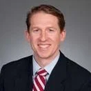 A headshot of a smiling man in a dark suit, white shirt, and striped tie, with short brown hair against a plain background.