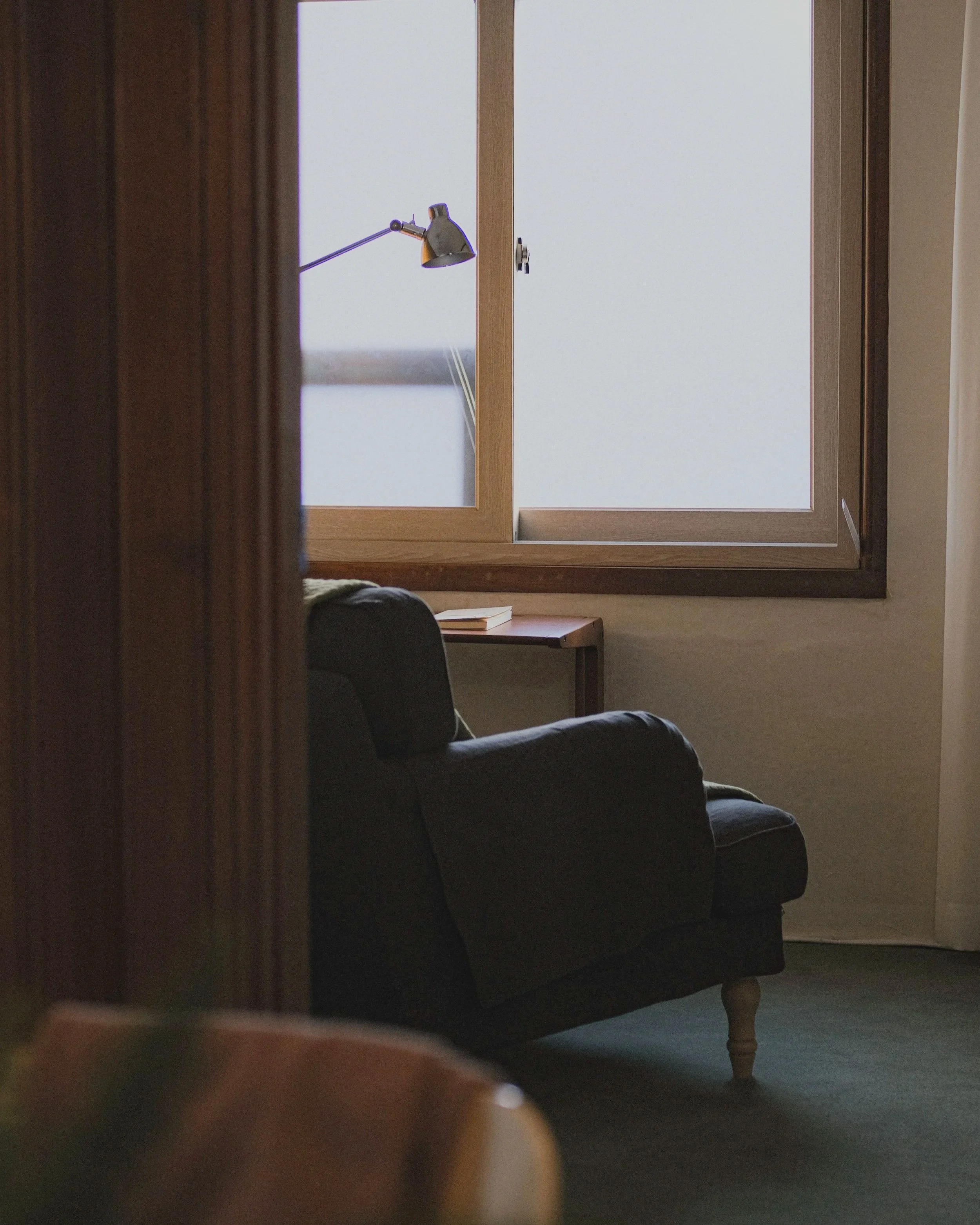A view through a partially open door showing a seating area with a sofa, a wooden side table with a closed book, a window with closed blinds, and a wall-mounted adjustable reading lamp.