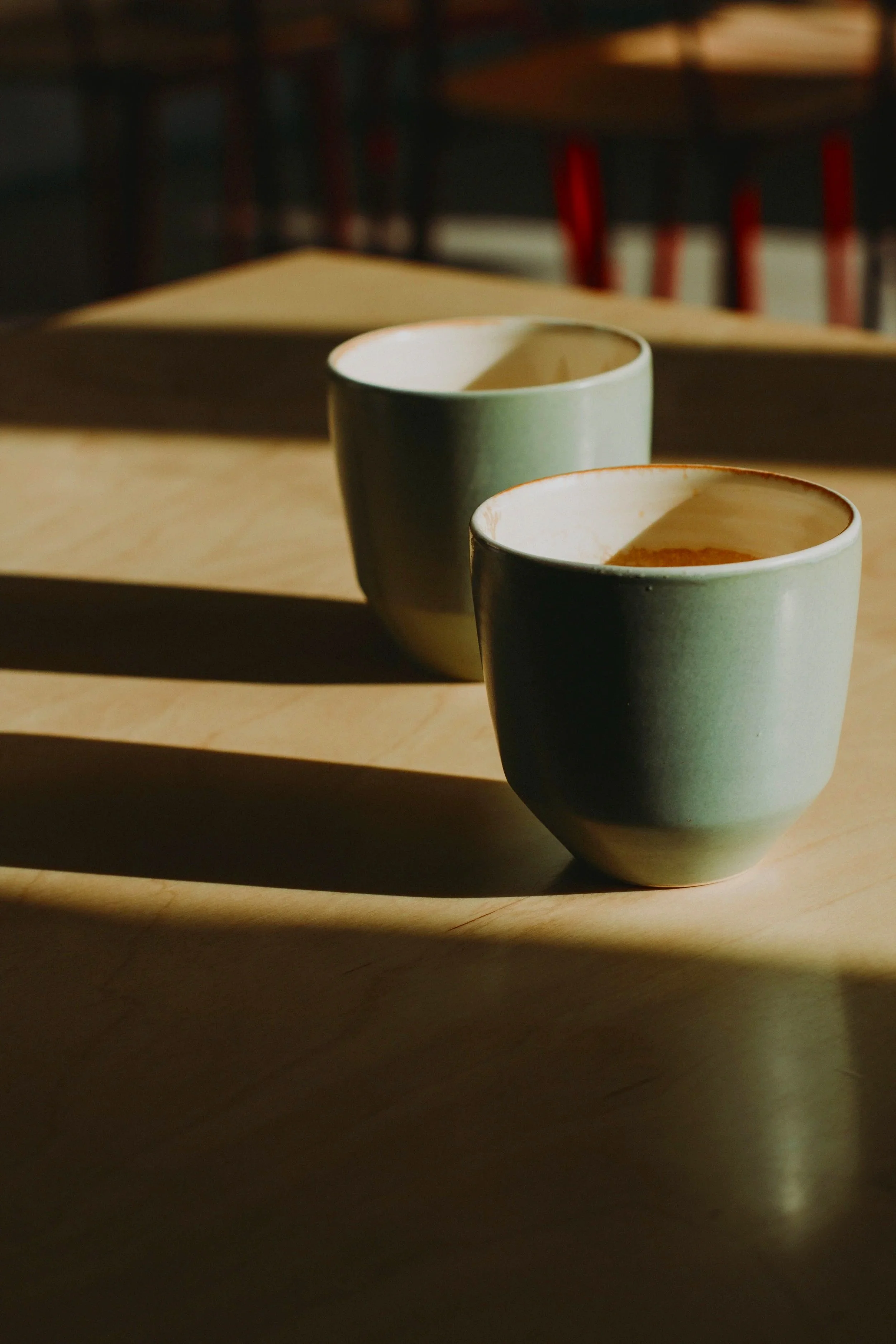 Two ceramic cups with warm beverages placed on a wooden surface, illuminated by sunlight, casting shadows.