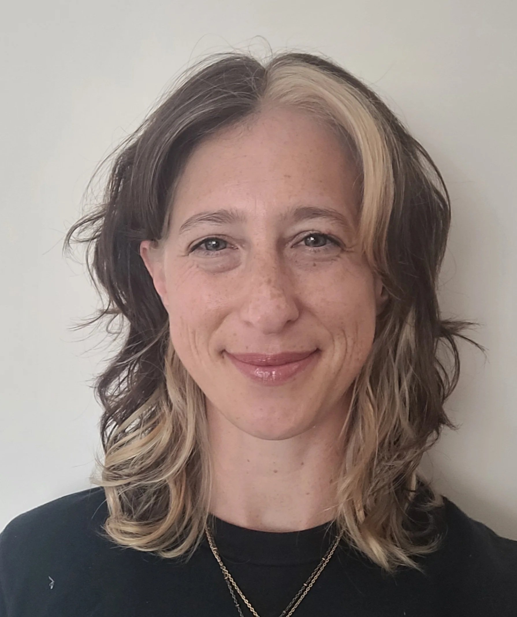 A woman with short hair, brown with blonde highlights, smiling against a plain white wall.