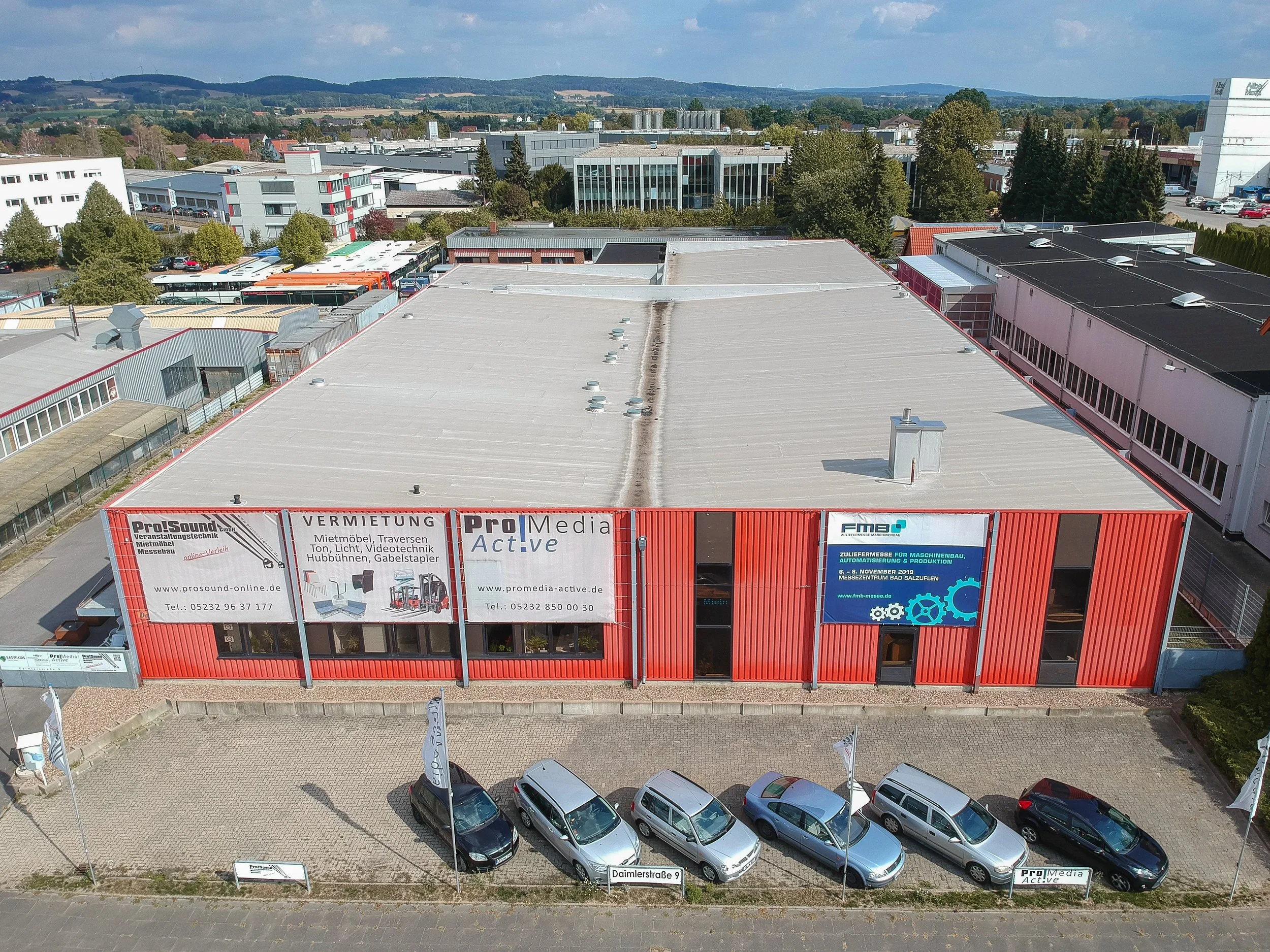 Luftaufnahme eines Industriegebäudes mit mehreren LKWs, Autos und Bäumen umgeben, im Hintergrund Hügel und bewölkter Himmel.