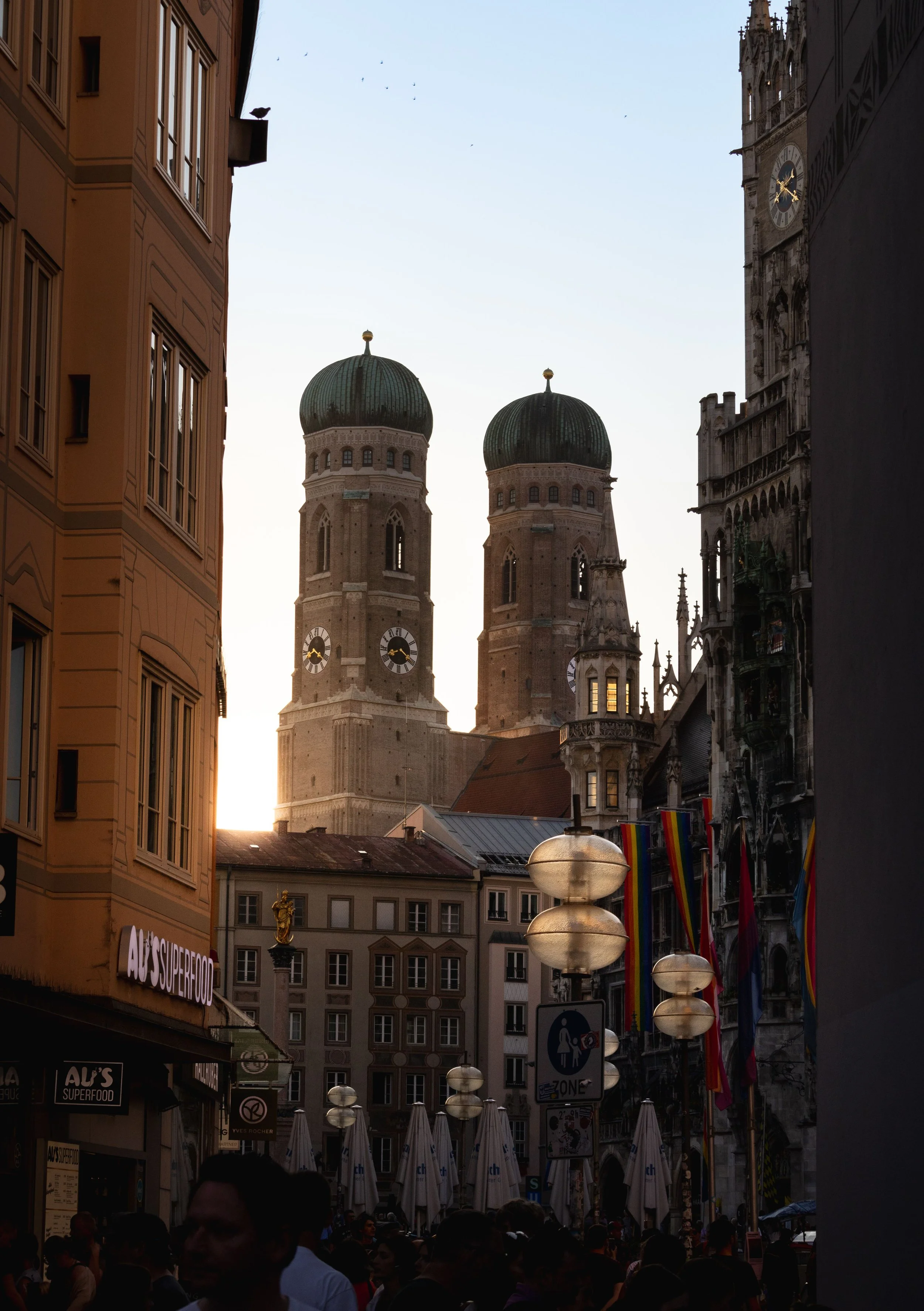 Blick durch eine belebte Straße in München mit historischen Gebäuden, Kirchtürmen im Hintergrund und Laternen, Menschen und bunten Fahnen.