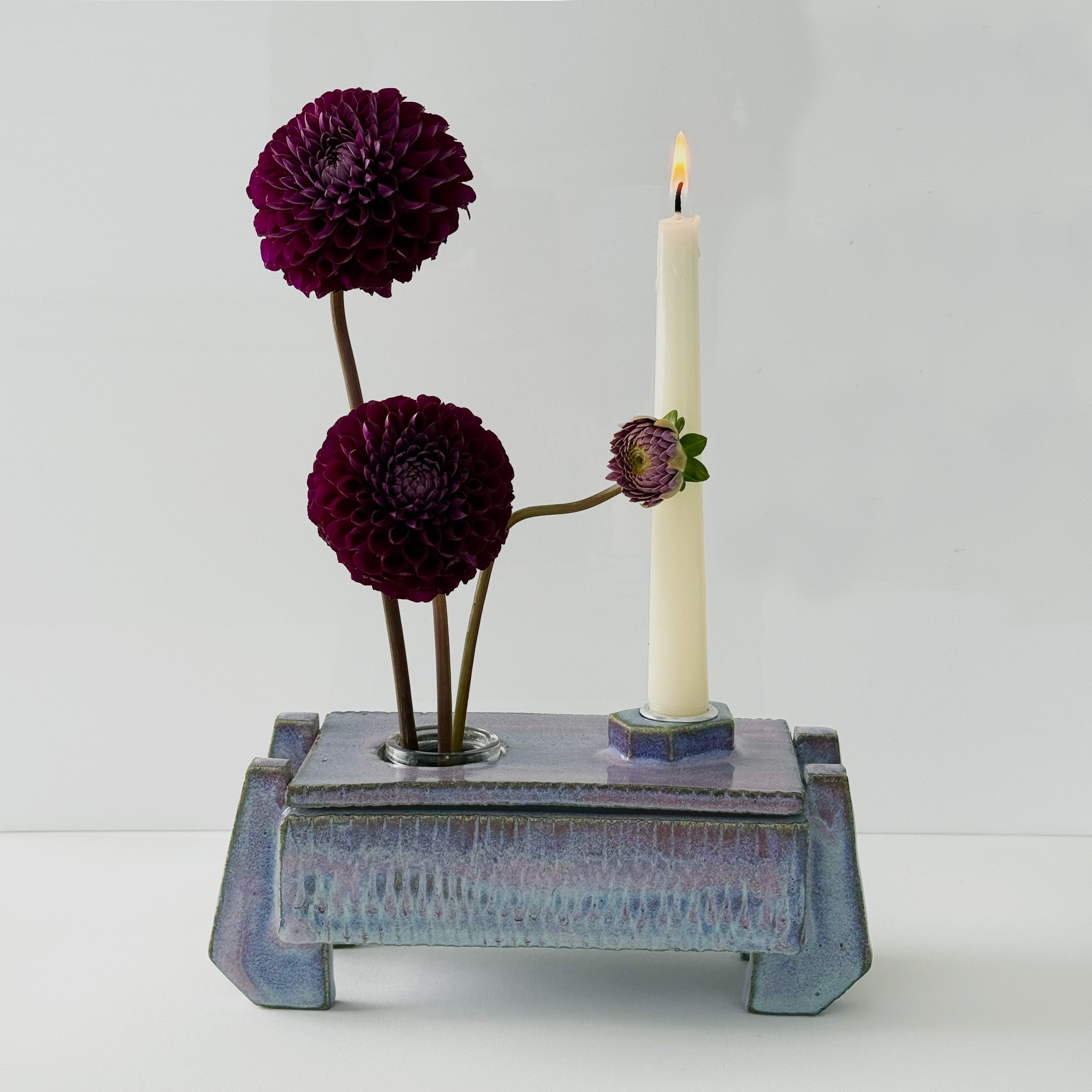 A still life composition featuring a purple dahlia flower, a lit white taper candle, and small purplish flowers, arranged on a small carved wooden stand with a plain white background.