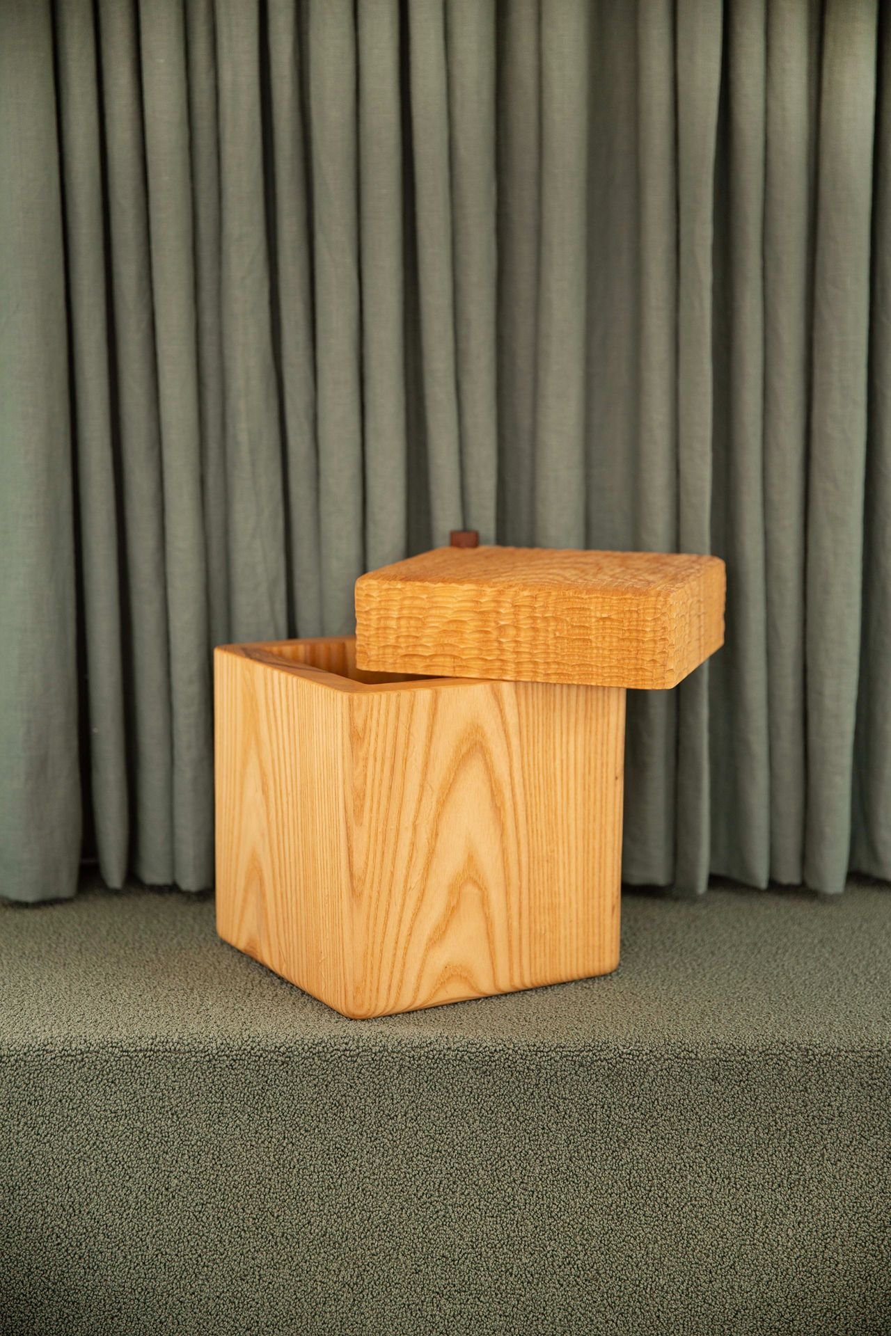 A wooden cube-shaped stool with a tilted rectangular seat, placed on a green carpet in front of a green curtained background.