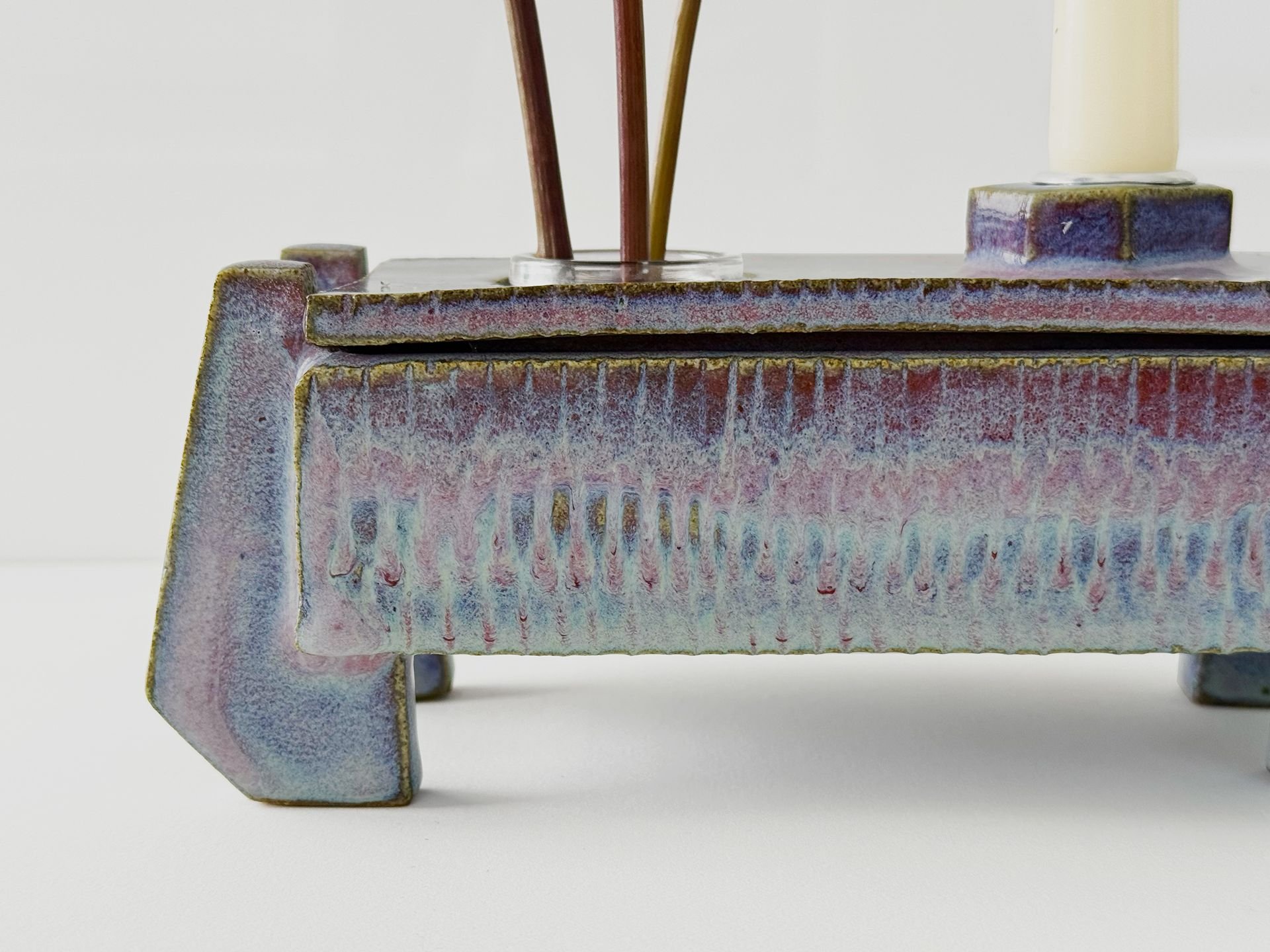 Close-up of a ceramic incense holder with a textured, multicolored glaze, holding several sticks of incense and a candle in a small ceramic holder on a white background.
