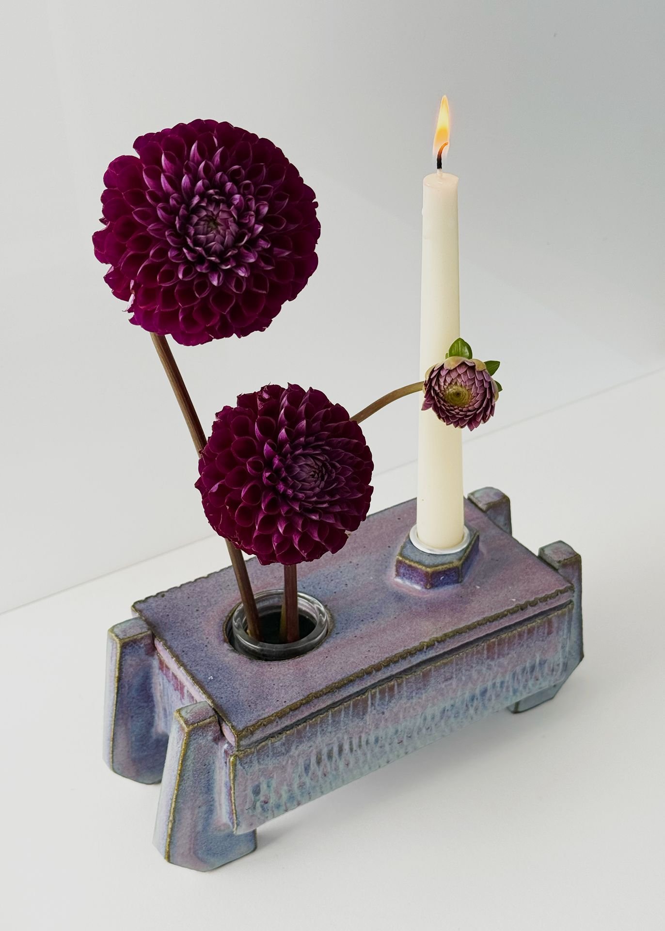 A ceramic vase with colorful glaze holds two purple dahlias, a small flower, and a white candle with a lit flame.